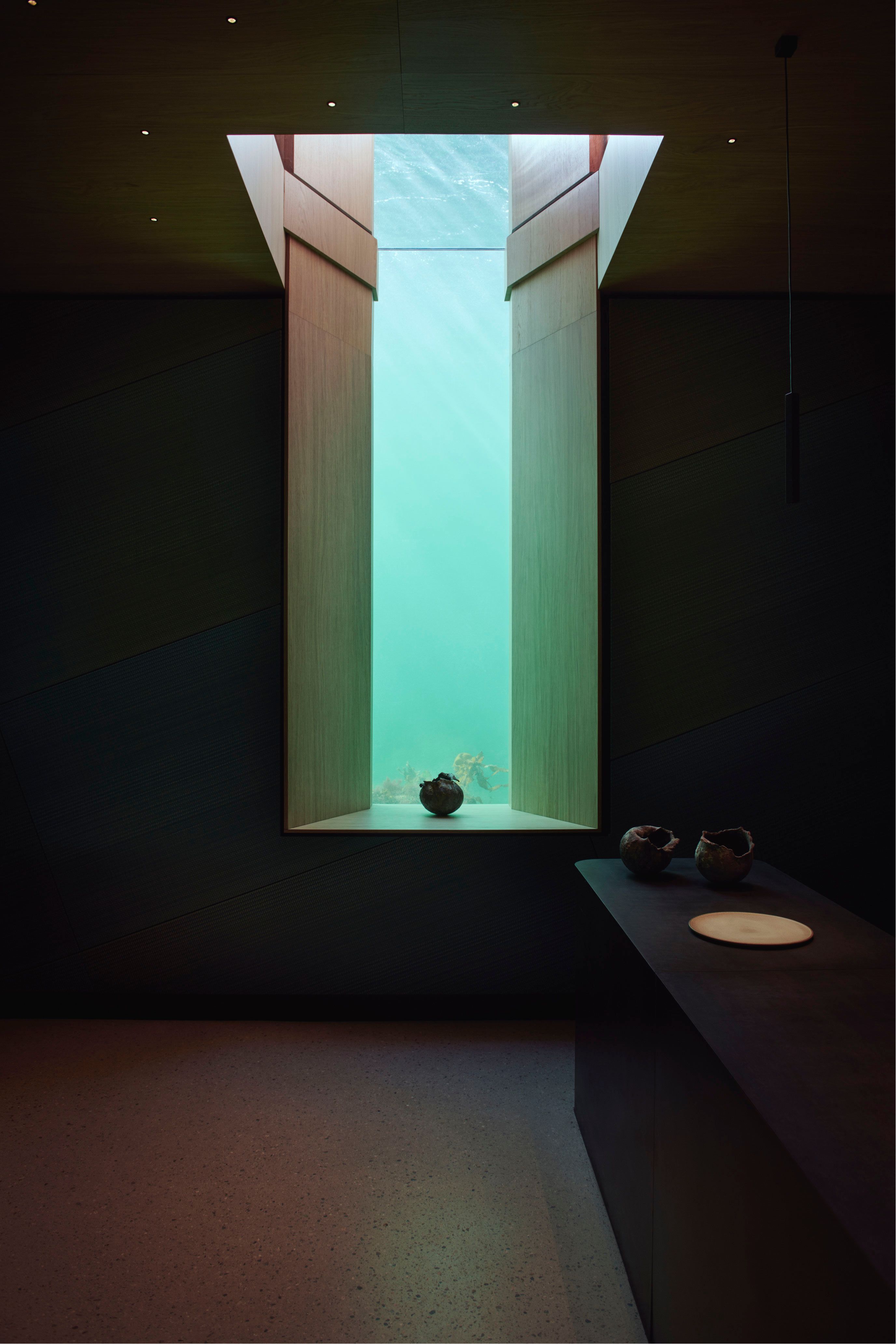 Narrow and tall window facing the water, turquoise-colored, algae at the bottom of the window. Dark room with a sideboard to the right.