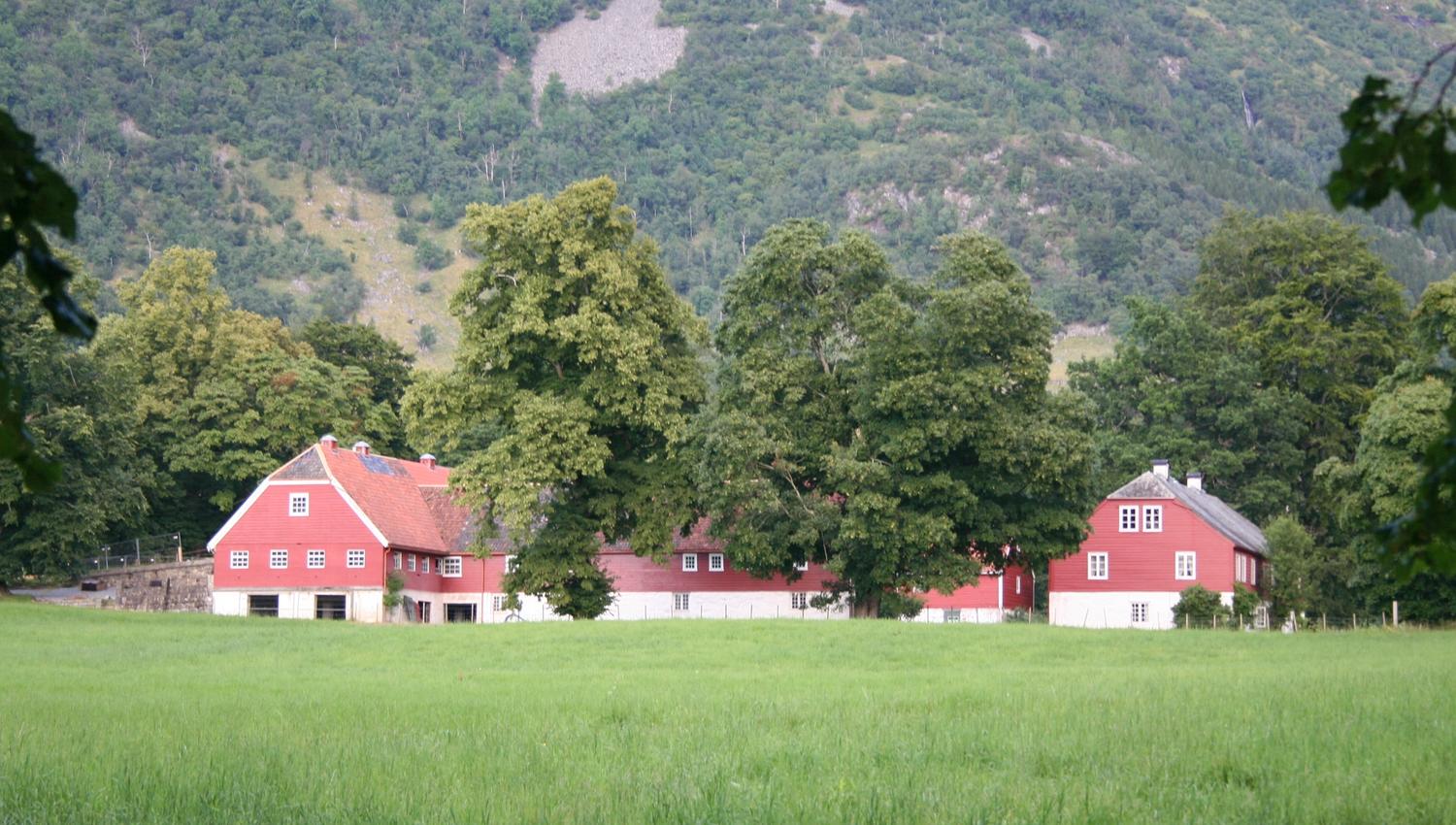 Dei raude uthusa ved Baroniet i Rosendal omgitt av grøne marker og majestetiske fjell.