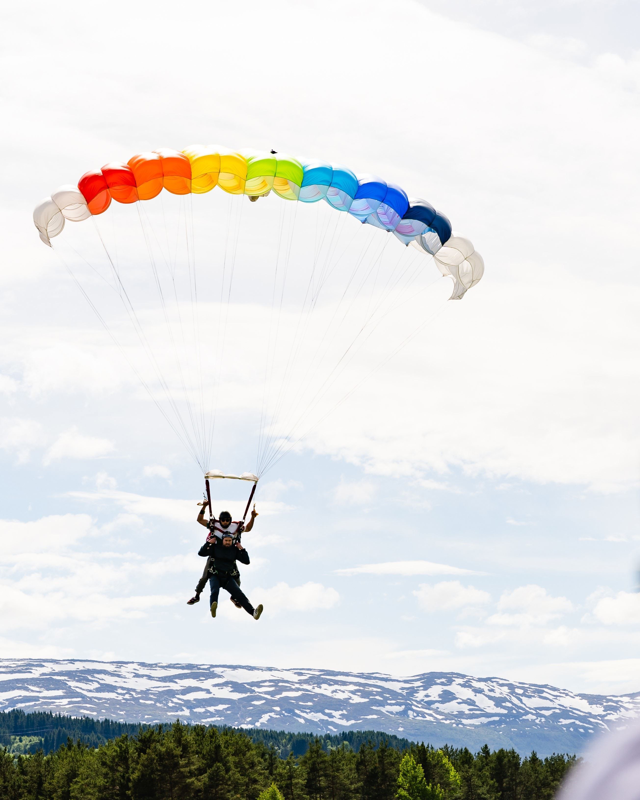 Tandem parachute descent under a vibrant, multi‑coloured canopy above verdant hills and snow‑dusted peaks.