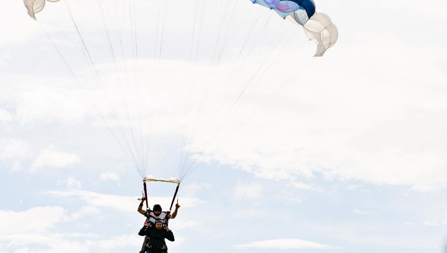 Tandem parachute descent under a vibrant, multi‑coloured canopy above verdant hills and snow‑dusted peaks.