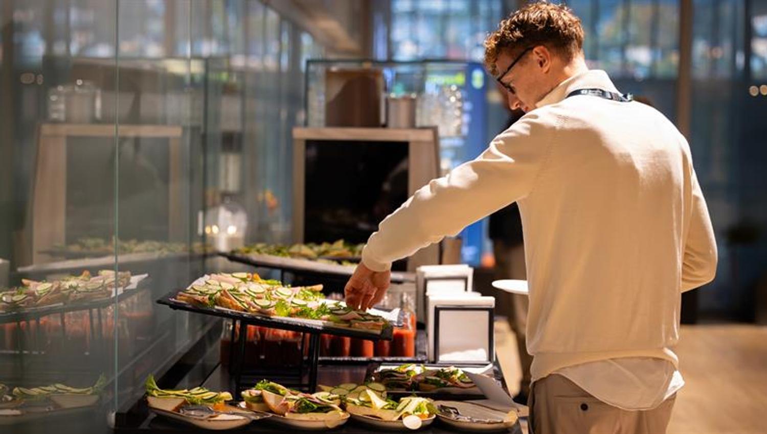 En mann med navneskilt forsyner seg av en buffet med smørbrød og salat i et lyst konferanseområde.