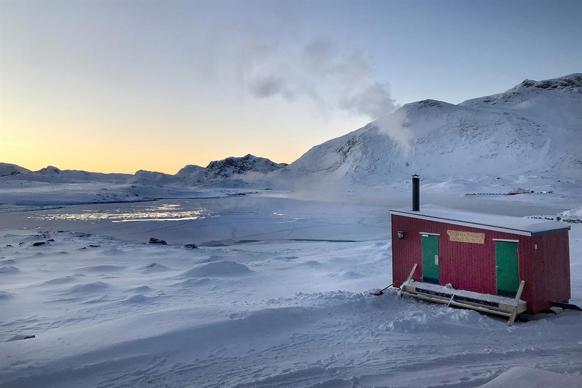 Liten rød utebadstue ved bredden av es islagt vann med fjell i bakgrunnen