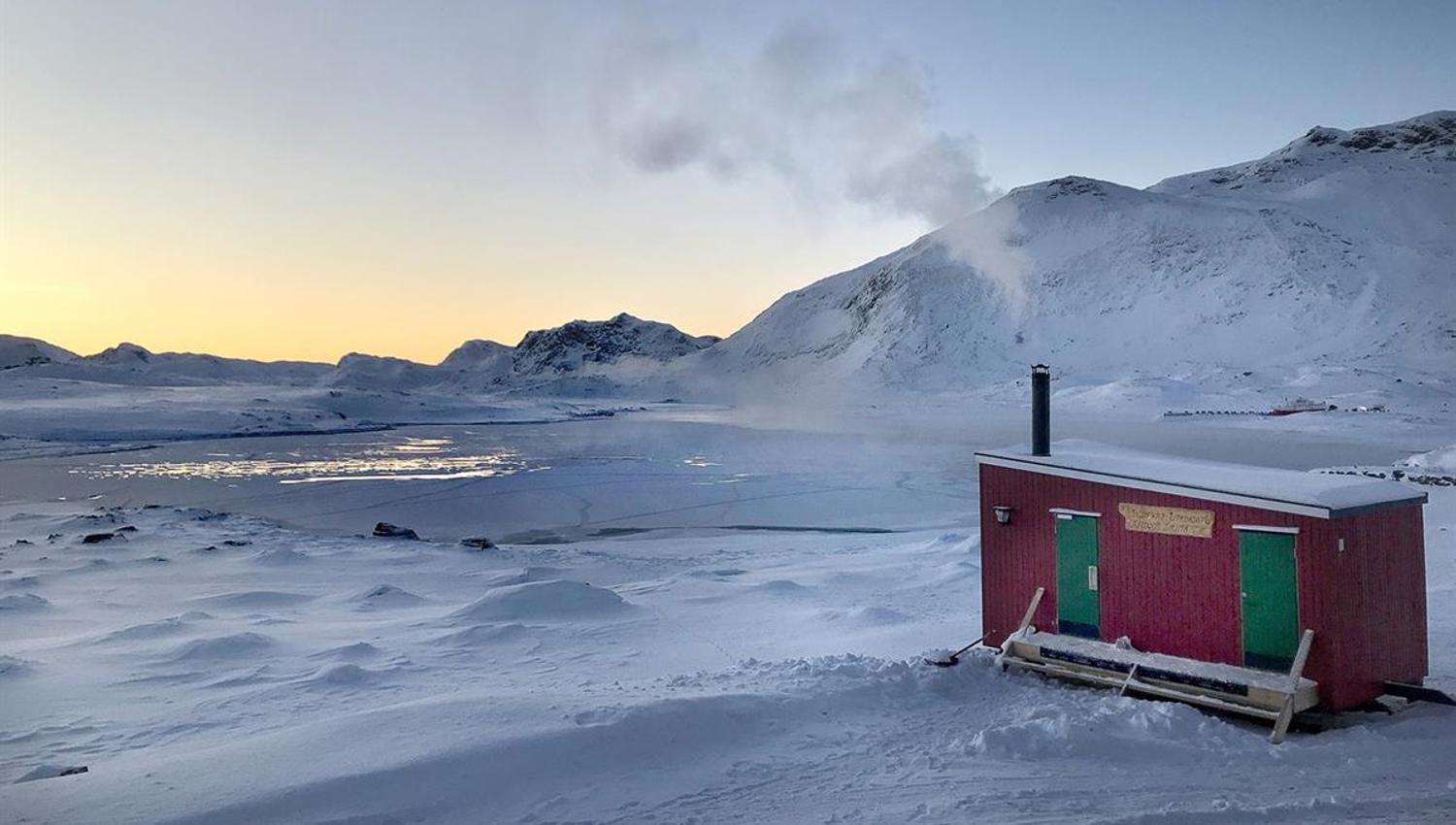 Liten rød utebadstue ved bredden av es islagt vann med fjell i bakgrunnen