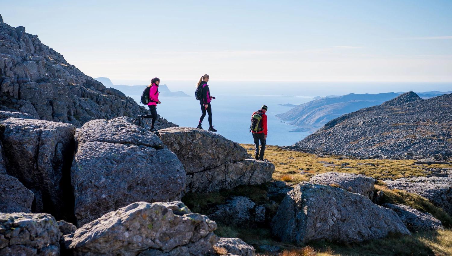 Three hikers with backpacks walk across rocky mountain terrain, overlooking a blue sea and distant islands..