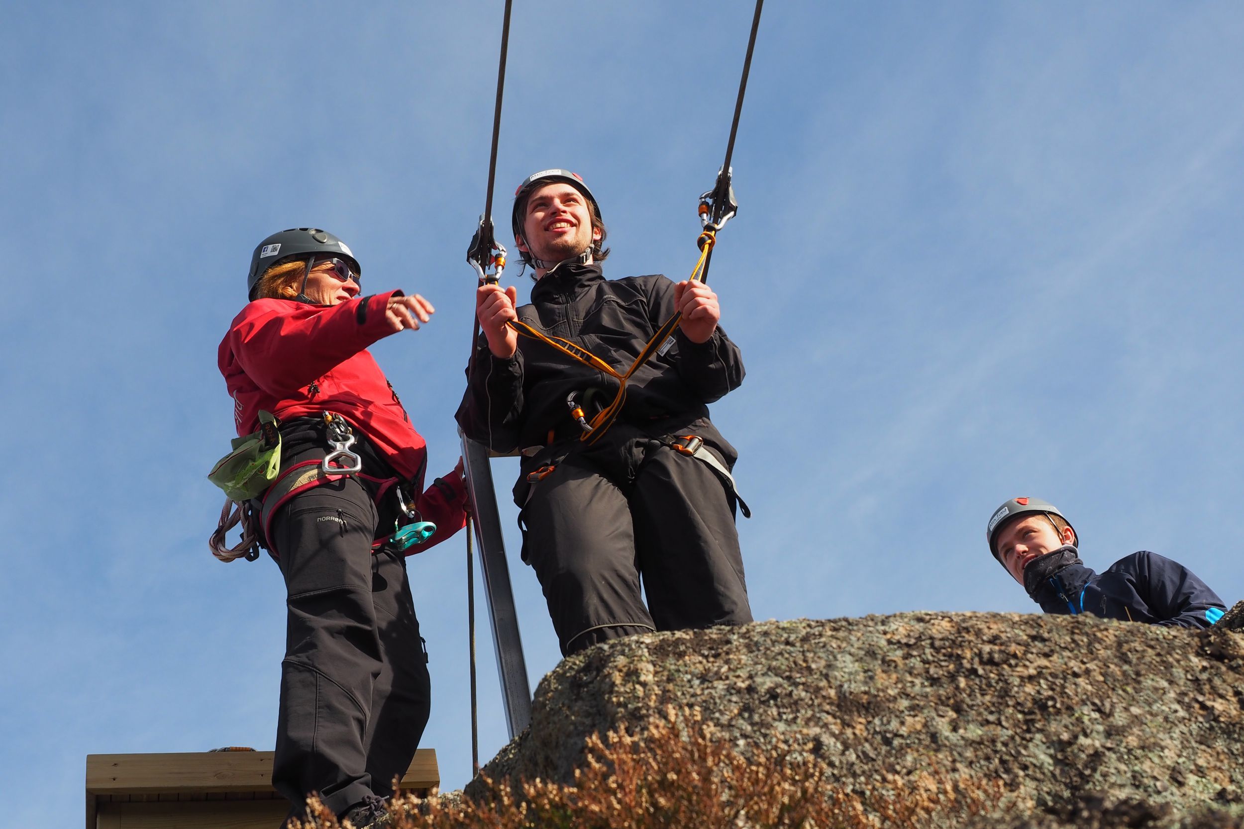 Berit instruerer en ung mann på vei ut i zipline. Bak står nestemann og venter på tur.