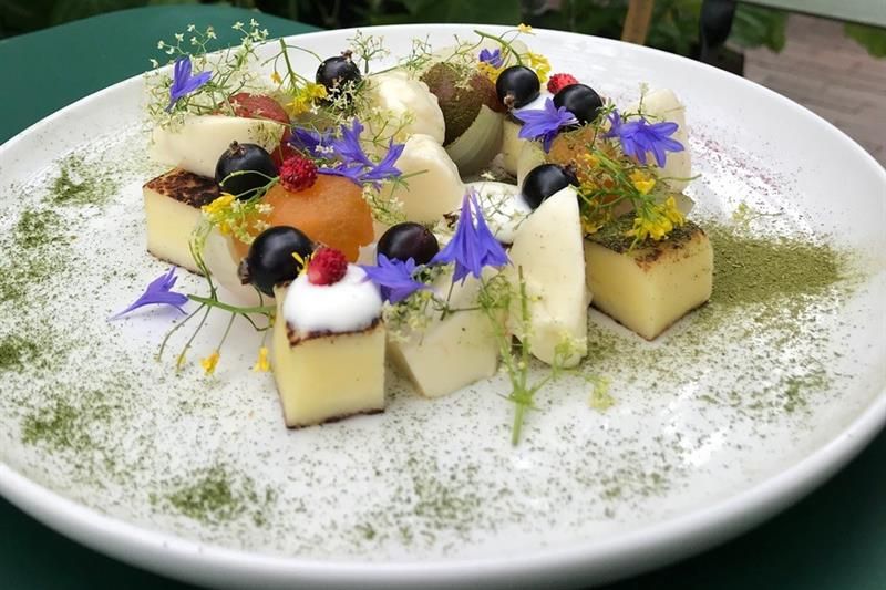 Et delikat dessertfat servert på en hvit tallerken, plassert på et grønt bord i et drivhus omgitt av grønne planter og ranker. Retten består av små terninger av ost eller flan, pyntet med spiselige blomster, urter, bær som solbær og markjordbær, samt små kuler av frukt eller sorbet. Tallerkenen er drysset med grønt pulver, sannsynligvis tørket urt. I bakgrunnen ses grønne planter og en grønn stol
