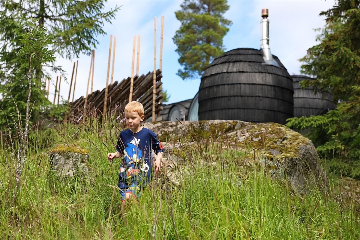 Child sitting in the grass in front of dark wooden cabin pods in the forest.