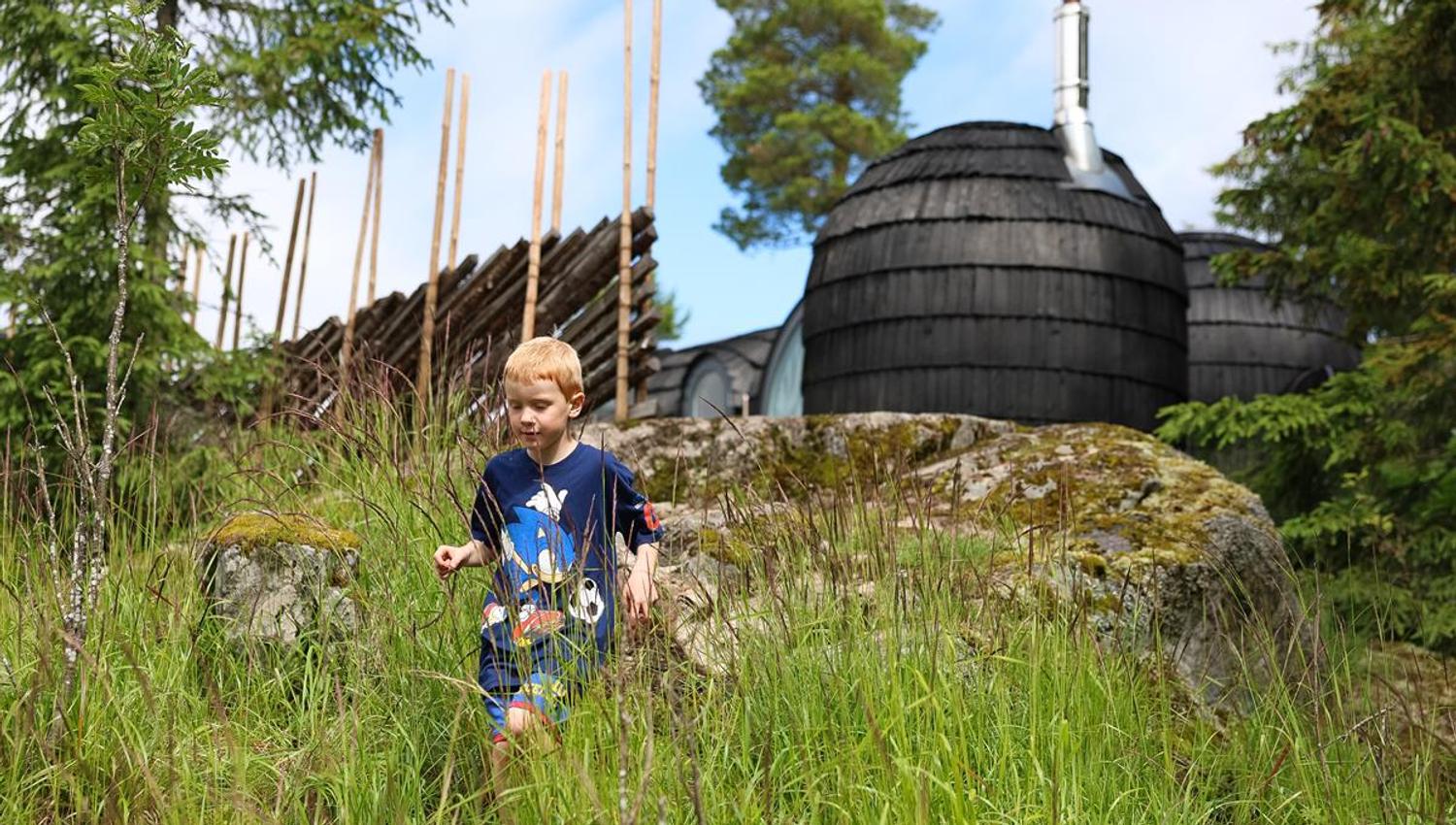 Child sitting in the grass in front of dark wooden cabin pods in the forest.