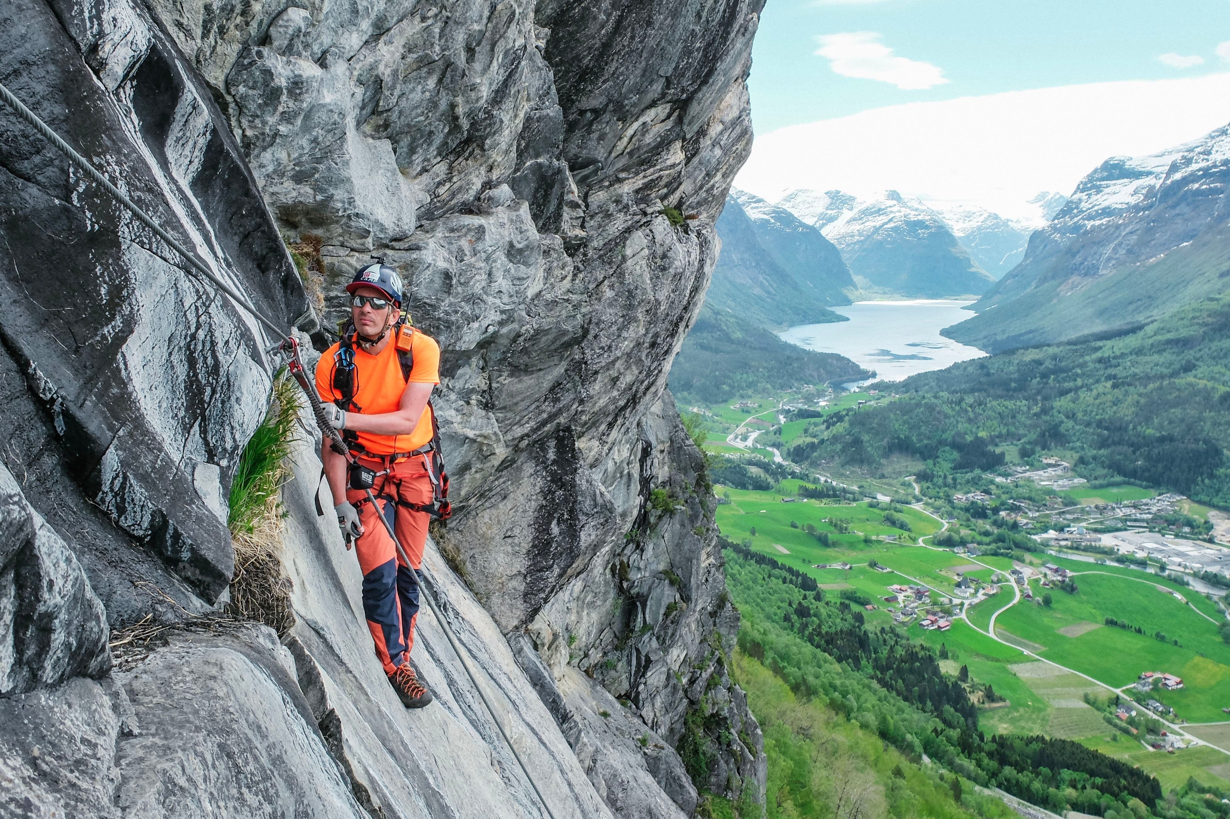 Ein klatrar i sele og hjelm ferdast langs ei bratt fjellhylle med tau, med dal, fjord og fjell i bakgrunnen..