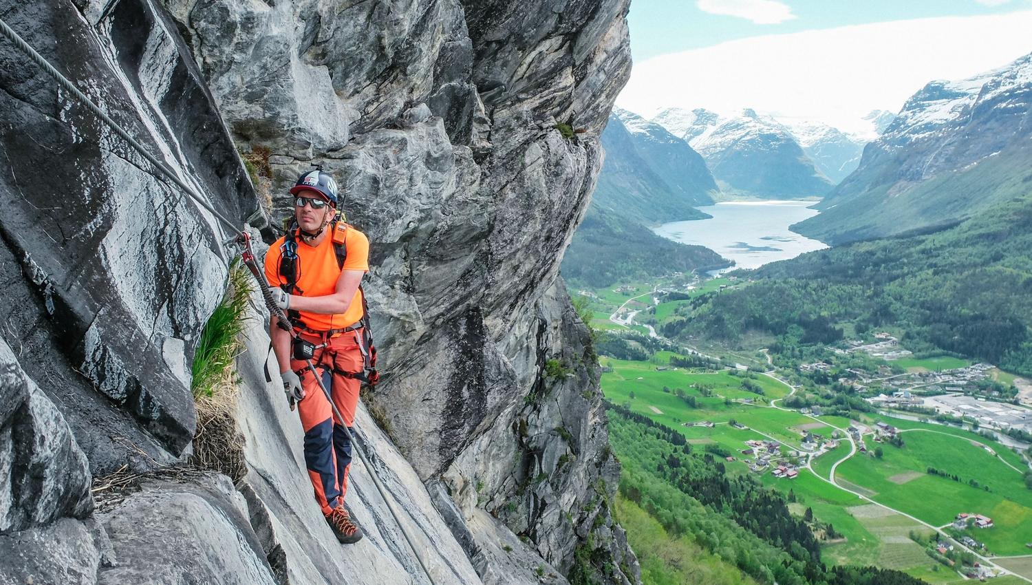 Ein klatrar i sele og hjelm ferdast langs ei bratt fjellhylle med tau, med dal, fjord og fjell i bakgrunnen..
