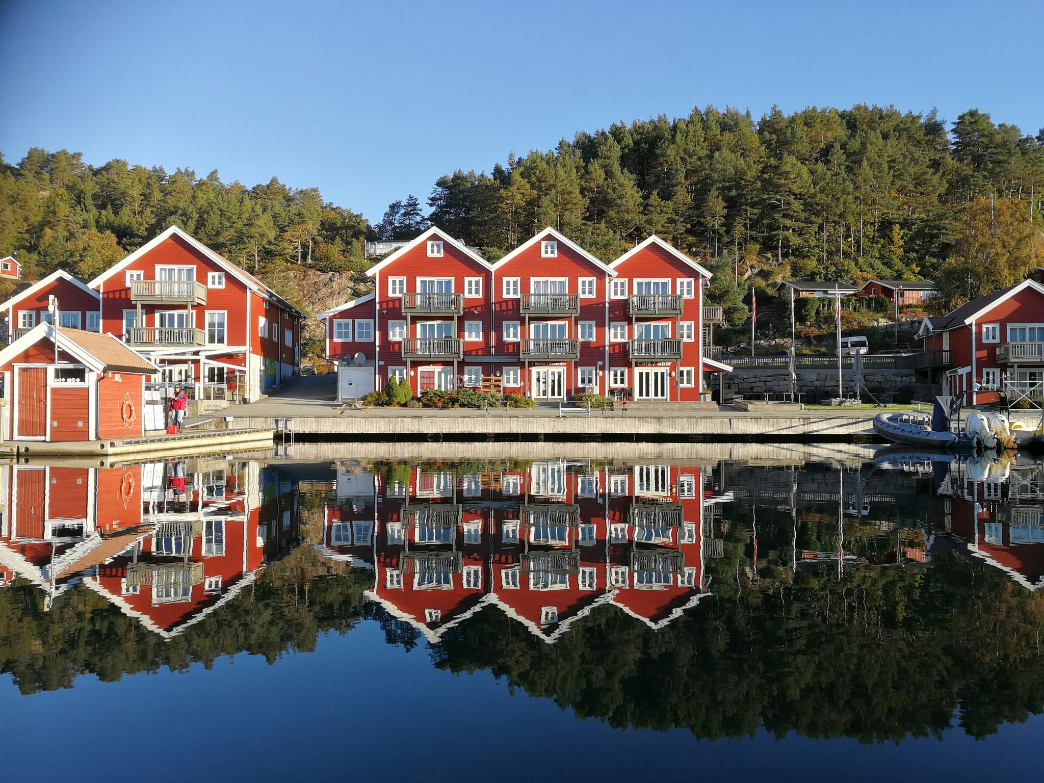 Tregde ferie, bilde tatt fra vannet inn mot brygga, røde bygninger med hvite karmer.