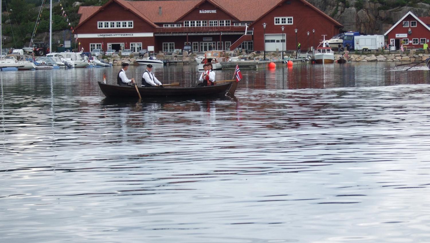 Liten trebåt på vannet med 2 menn og 2 kvinner i bunader, og det norske flagget på tuppen av båten foran den røde Lindesnes Hytteservice bygningen.