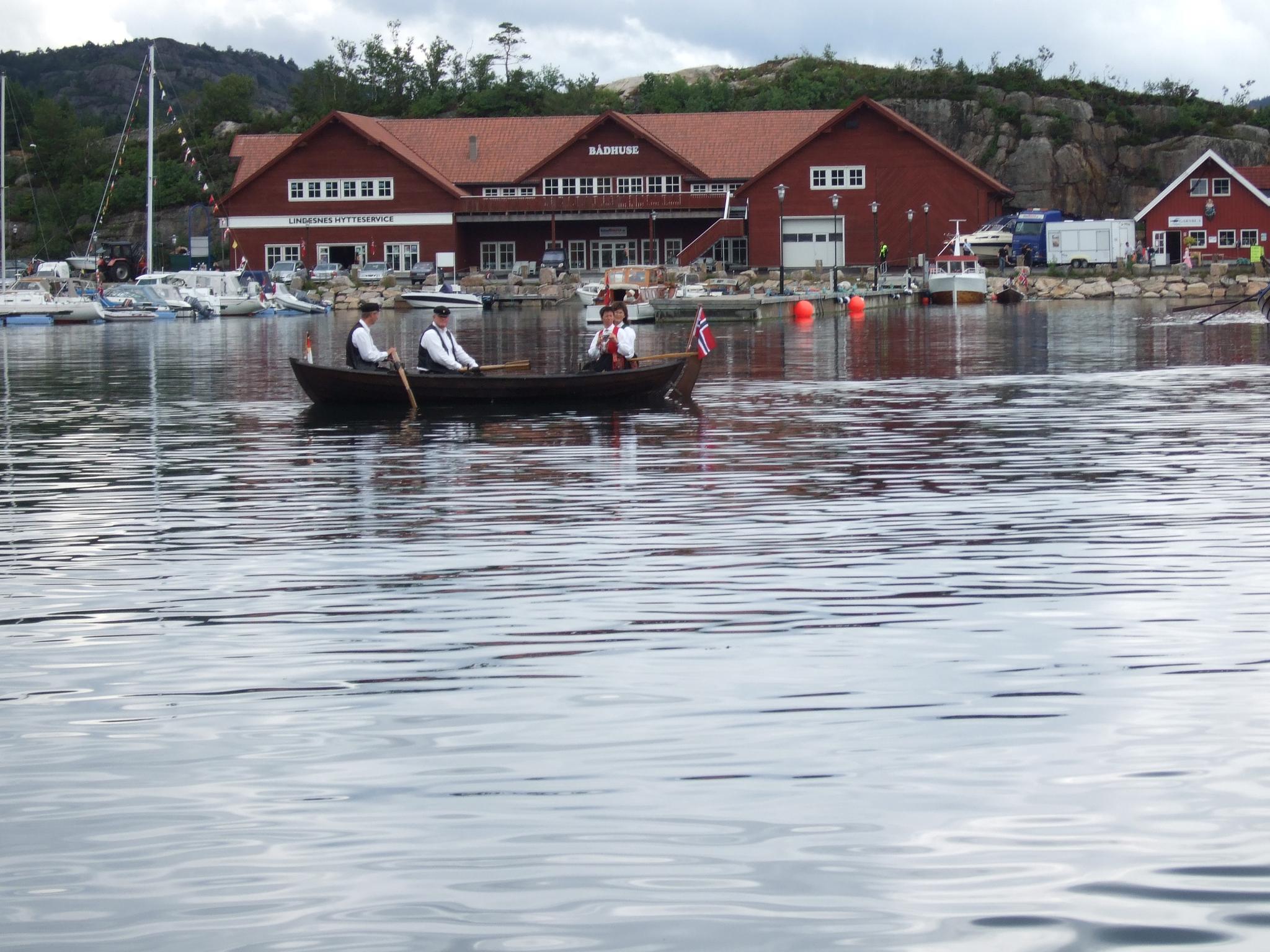 Liten trebåt på vannet med 2 menn og 2 kvinner i bunader, og det norske flagget på tuppen av båten foran den røde Lindesnes Hytteservice bygningen.