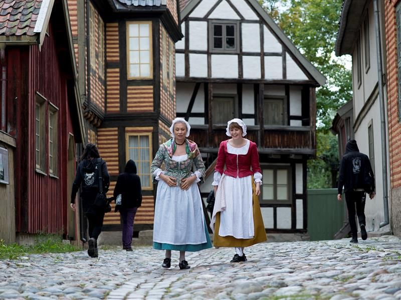 To kvinner i tradisjonelle klær går forbi gamle hus. Norsk Folkemuseum
