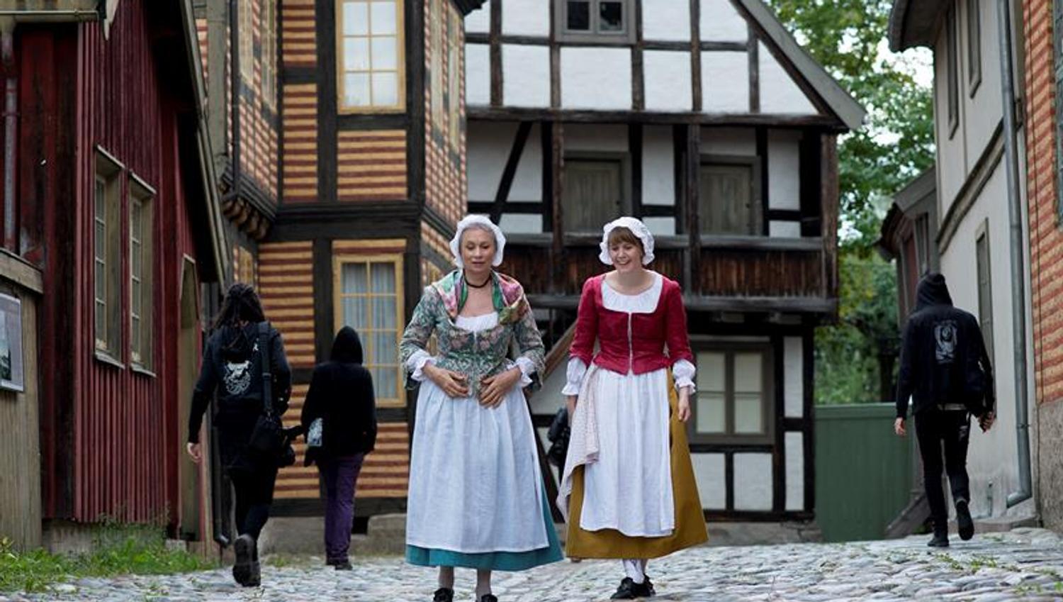 To kvinner i tradisjonelle klær går forbi gamle hus. Norsk Folkemuseum
