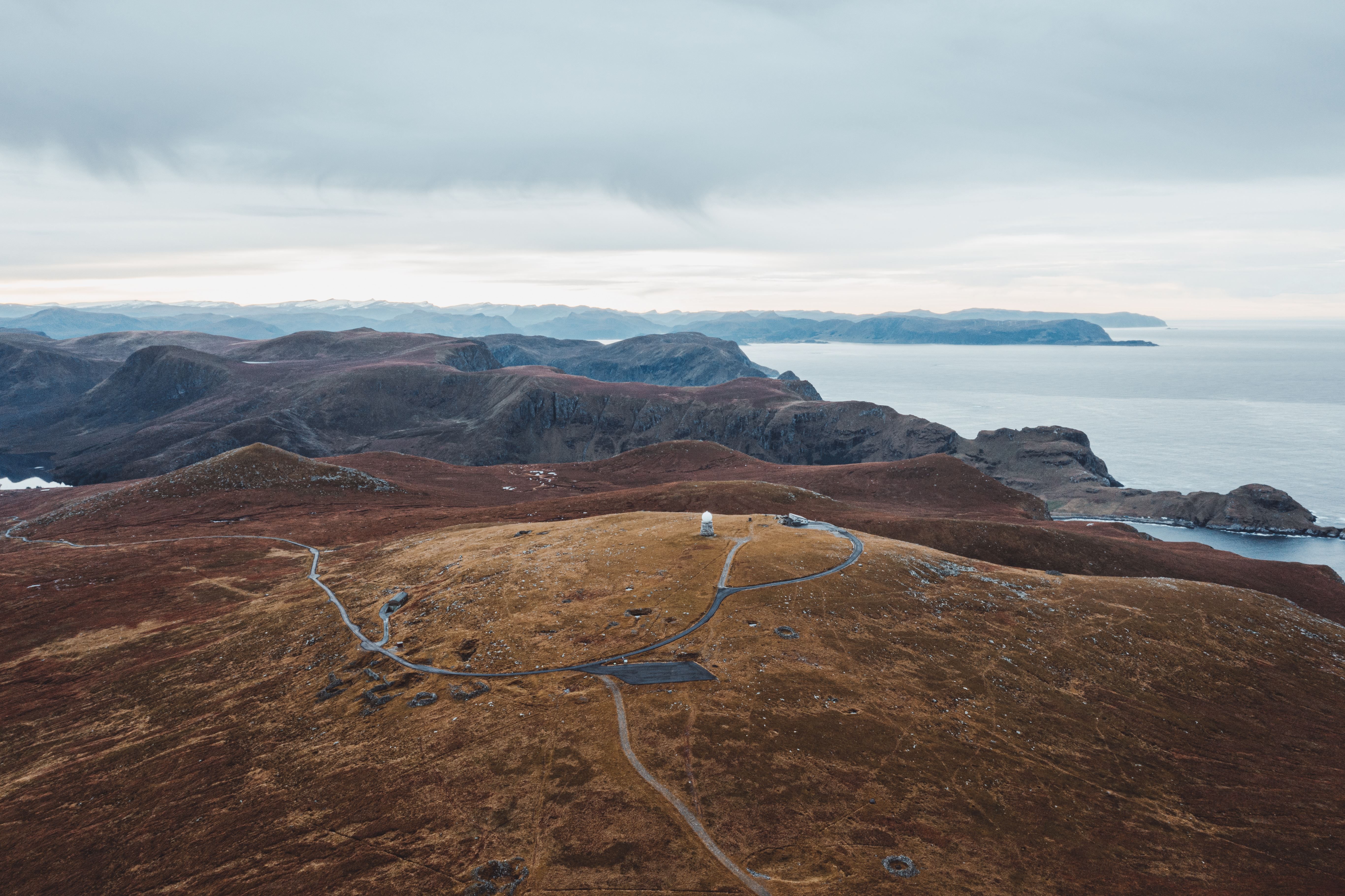 Vidt landskap med brun ås i forgrunnen, svingande veg og lite tårn, fjellryggar og hav under grå skyer..