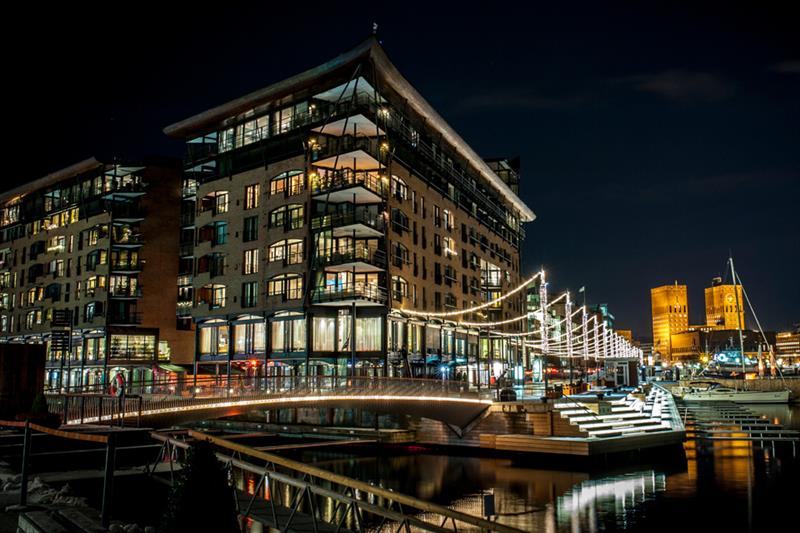Kveldsbilde av brygga med lysdekket bro og promenade. Rådhuset i bakgrunnen.