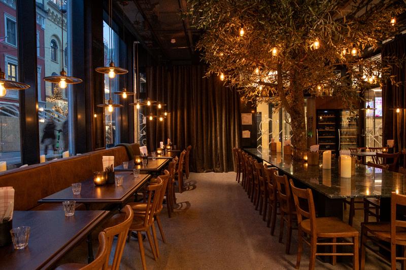 A restaurant with a modern glass façade and a warm, atmospheric interior featuring wooden tables, hanging lights, and set tables along the windows.