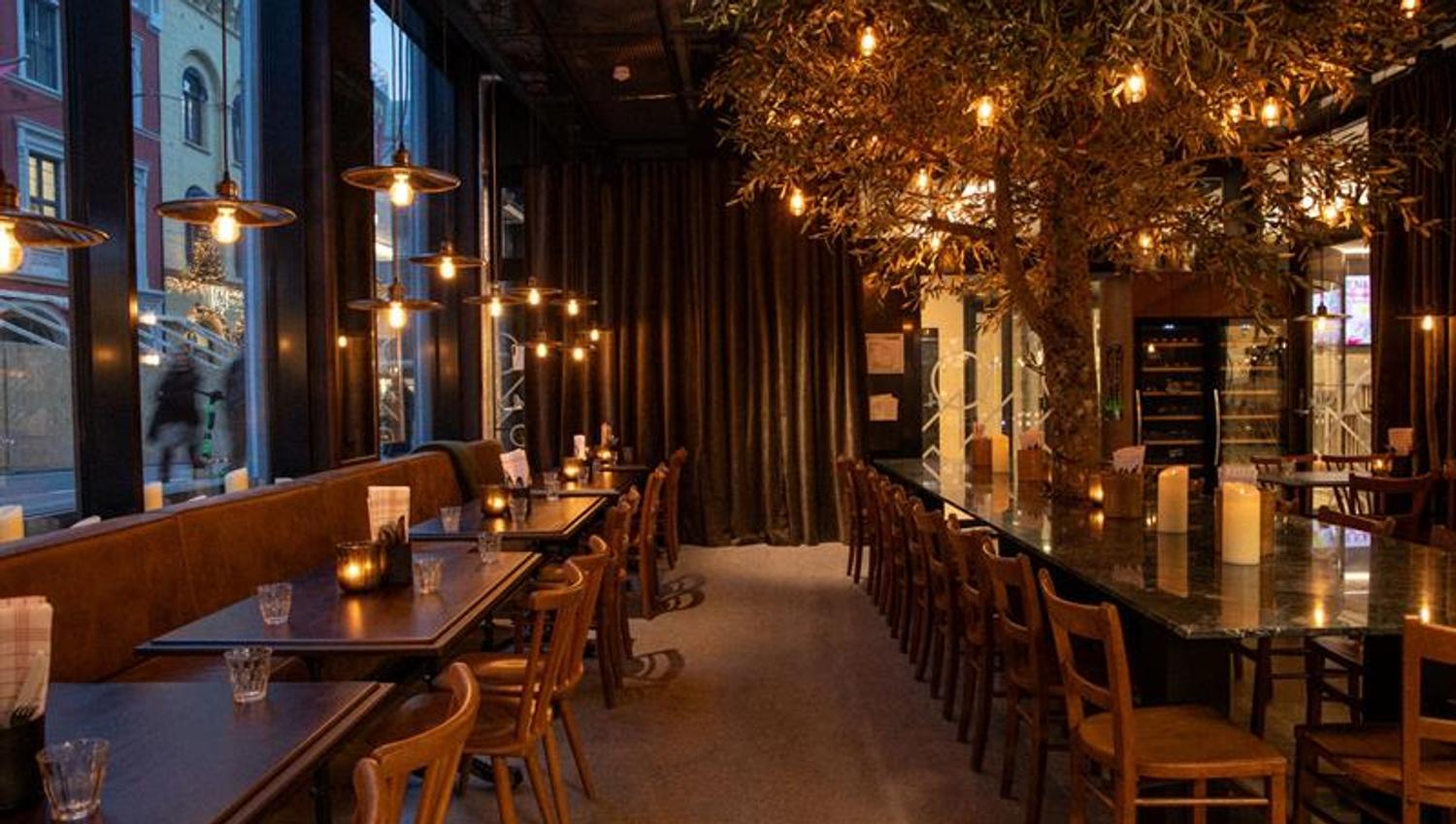 A restaurant with a modern glass façade and a warm, atmospheric interior featuring wooden tables, hanging lights, and set tables along the windows.