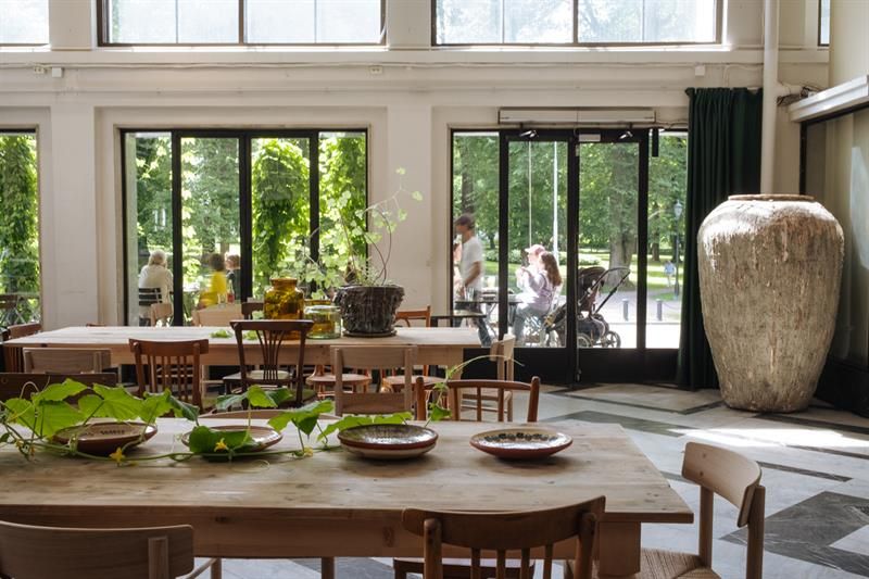 Two solid wood square dining tables set with bowls and green plants, wooden chairs around the tables, a large jar, and large windows overlooking the o