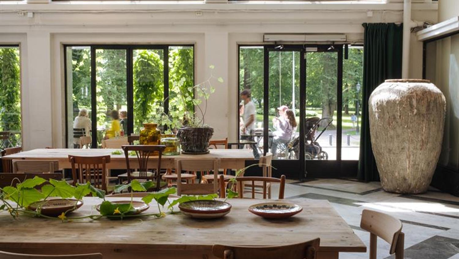 Two solid wood square dining tables set with bowls and green plants, wooden chairs around the tables, a large jar, and large windows overlooking the o