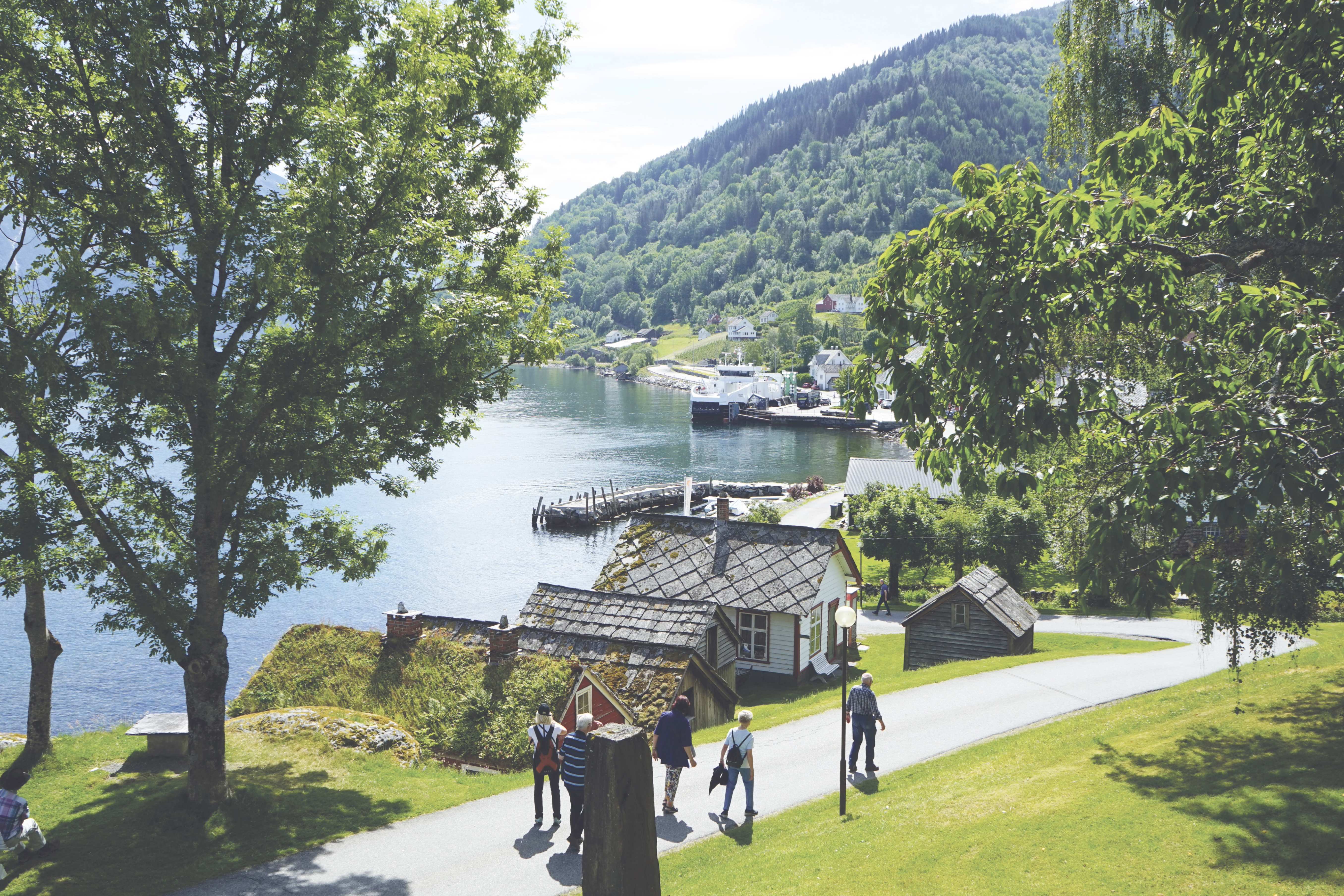 Ein gruppe mennesker går på ein veg med grønt gras og naust på venstre side. I bakgrunnen kan ein sjå ei ferge og vann og fjell.