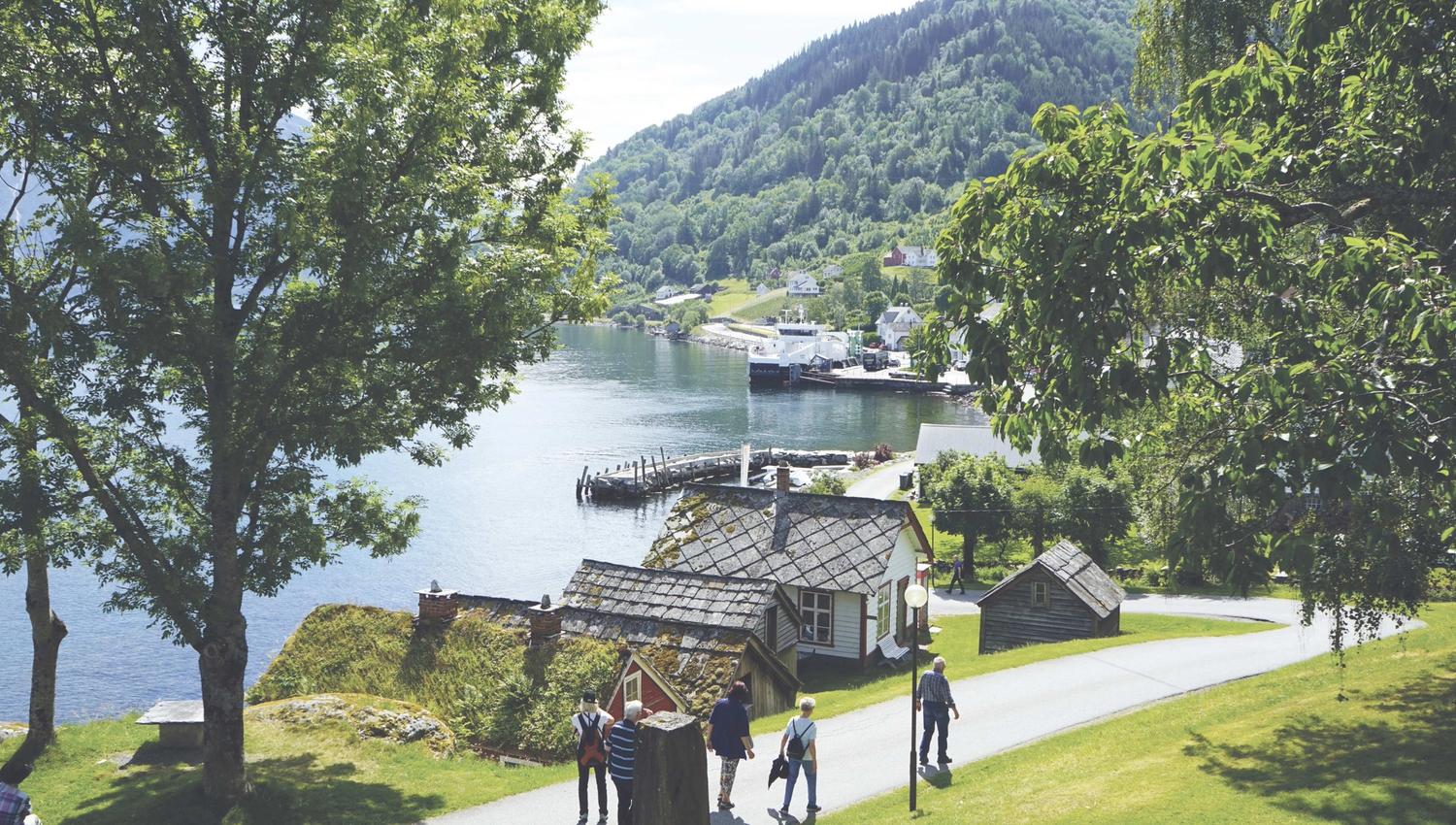 Ein gruppe mennesker går på ein veg med grønt gras og naust på venstre side. I bakgrunnen kan ein sjå ei ferge og vann og fjell.