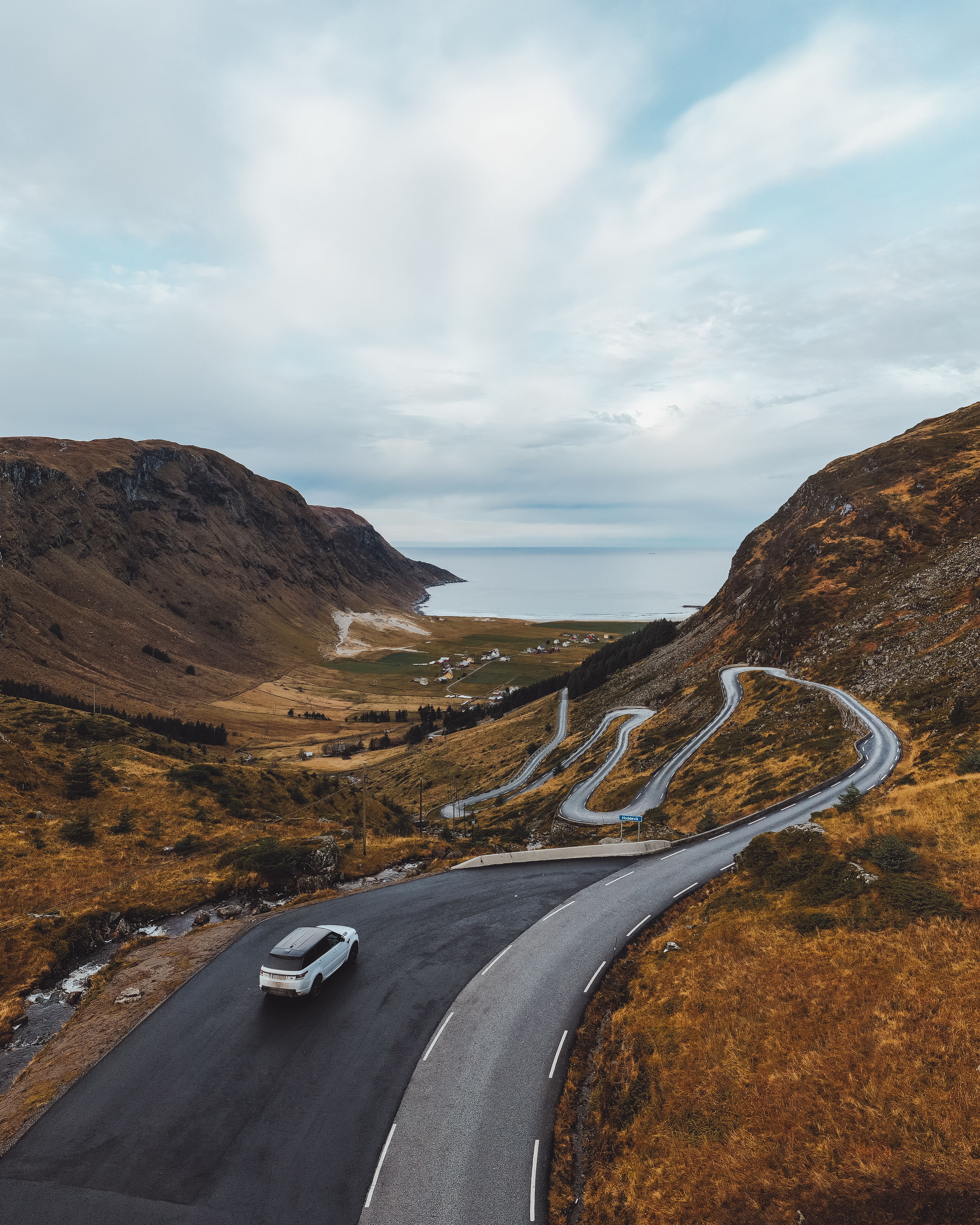 A car drives on a winding mountain road above a valley with small houses, leading toward a calm sea under clouds..