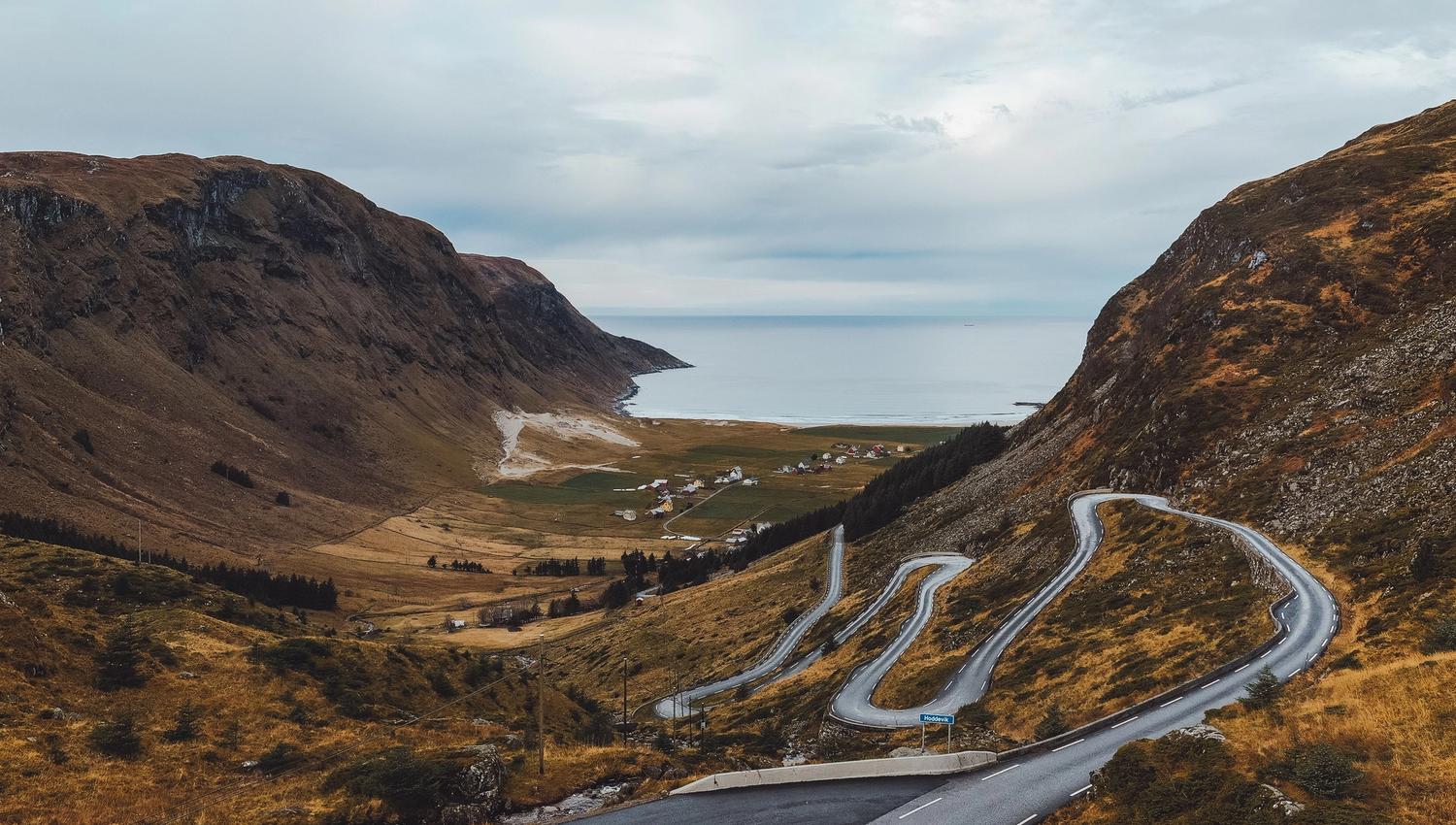 A car drives on a winding mountain road above a valley with small houses, leading toward a calm sea under clouds..