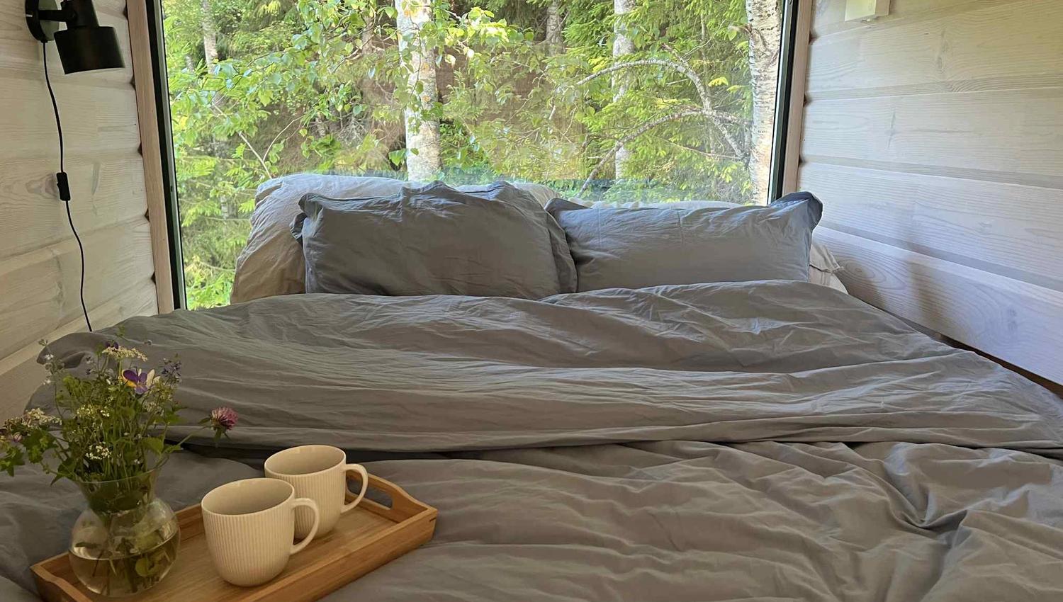 Bed in front of a large window with a view of nature, with a tray holding two coffee cups on it.
