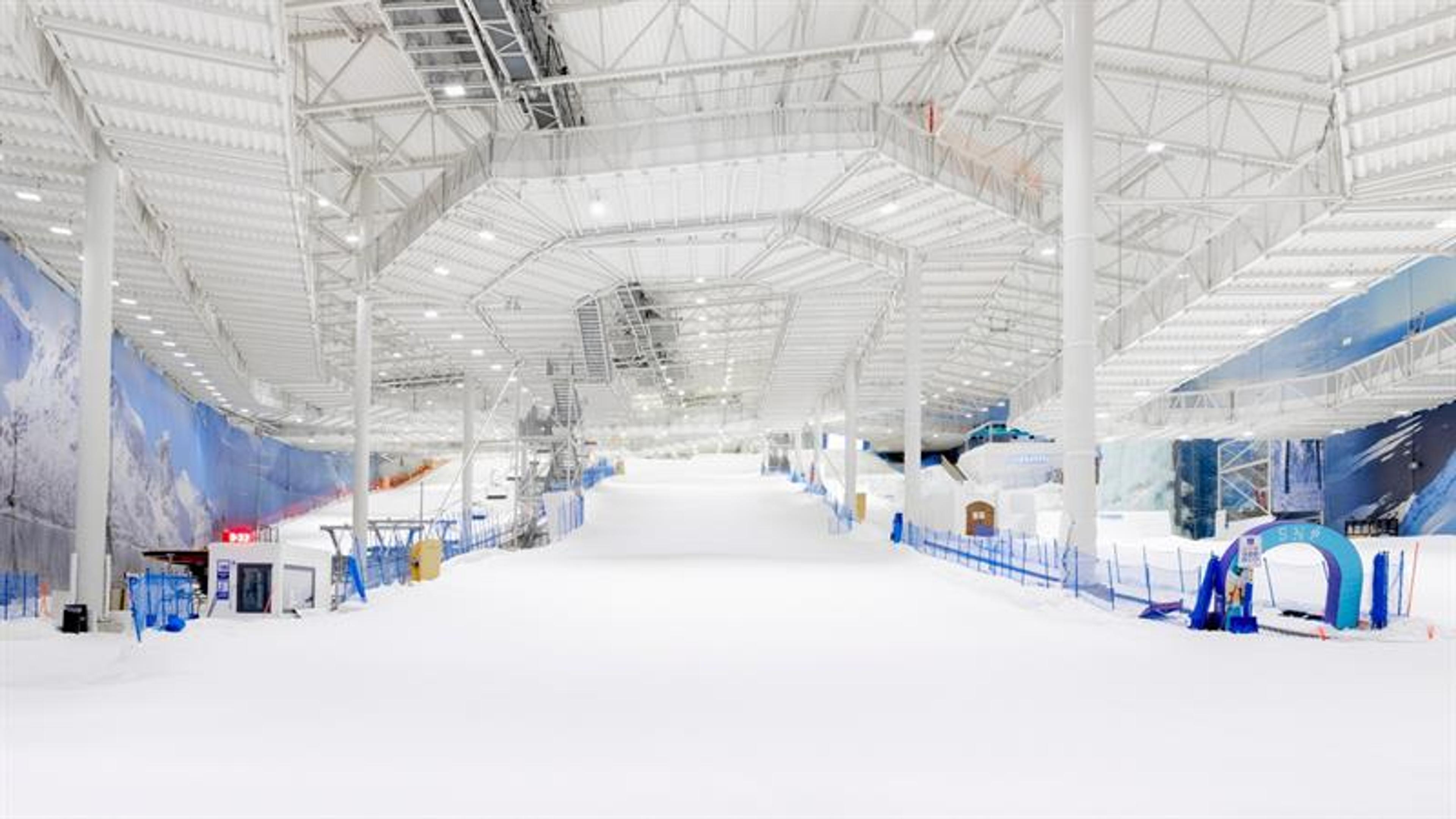 Indoor ski facility with a snow-covered slope, ski lift and high ceiling inside a large, modern hall.