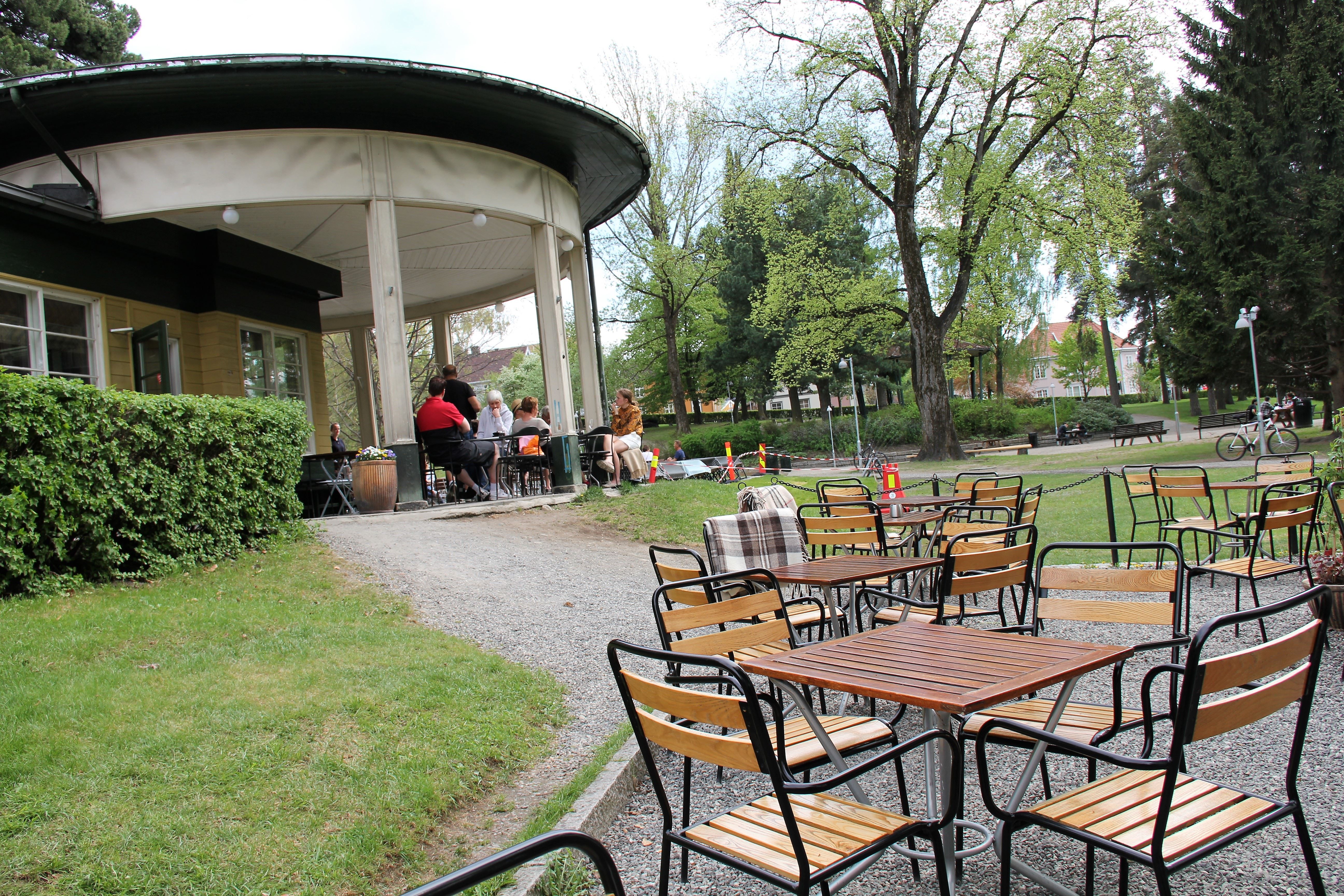 Uteservering med tomme bord og stoler foran en rund paviljong, med noen gjester under tak i en grønn park..