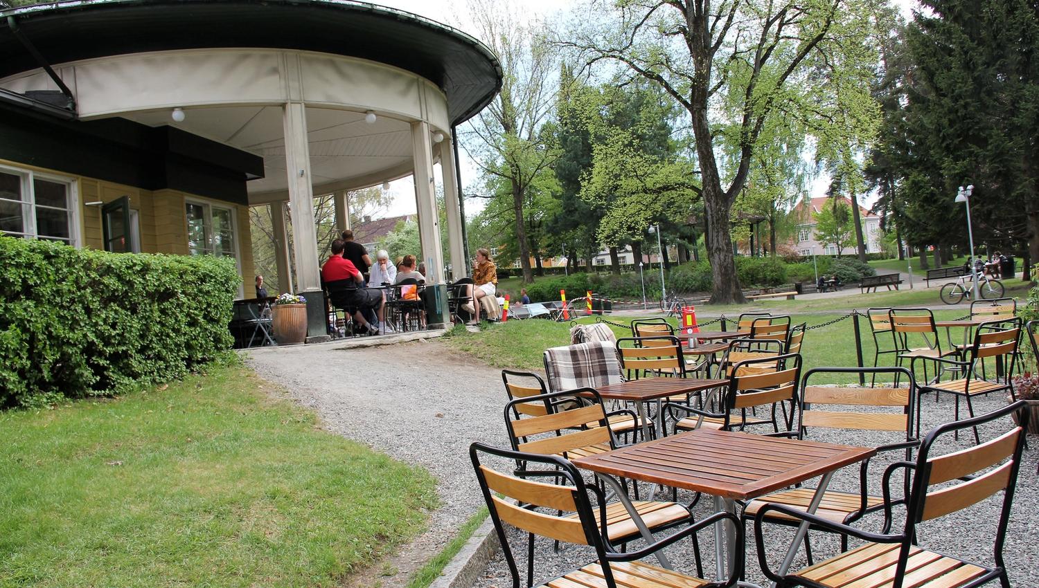 Uteservering med tomme bord og stoler foran en rund paviljong, med noen gjester under tak i en grønn park..