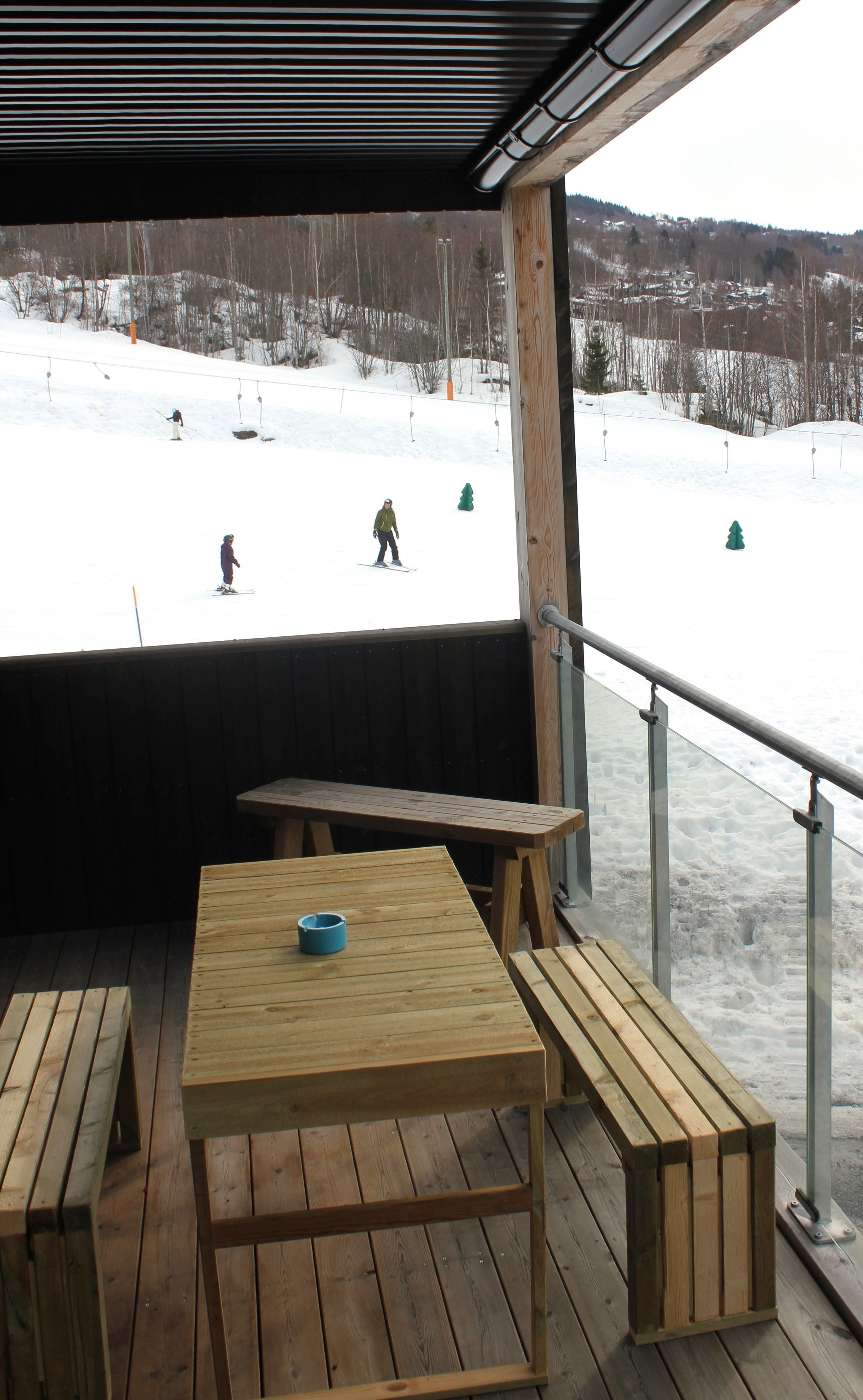 A table and benches on a terrace with snow and people skiing.