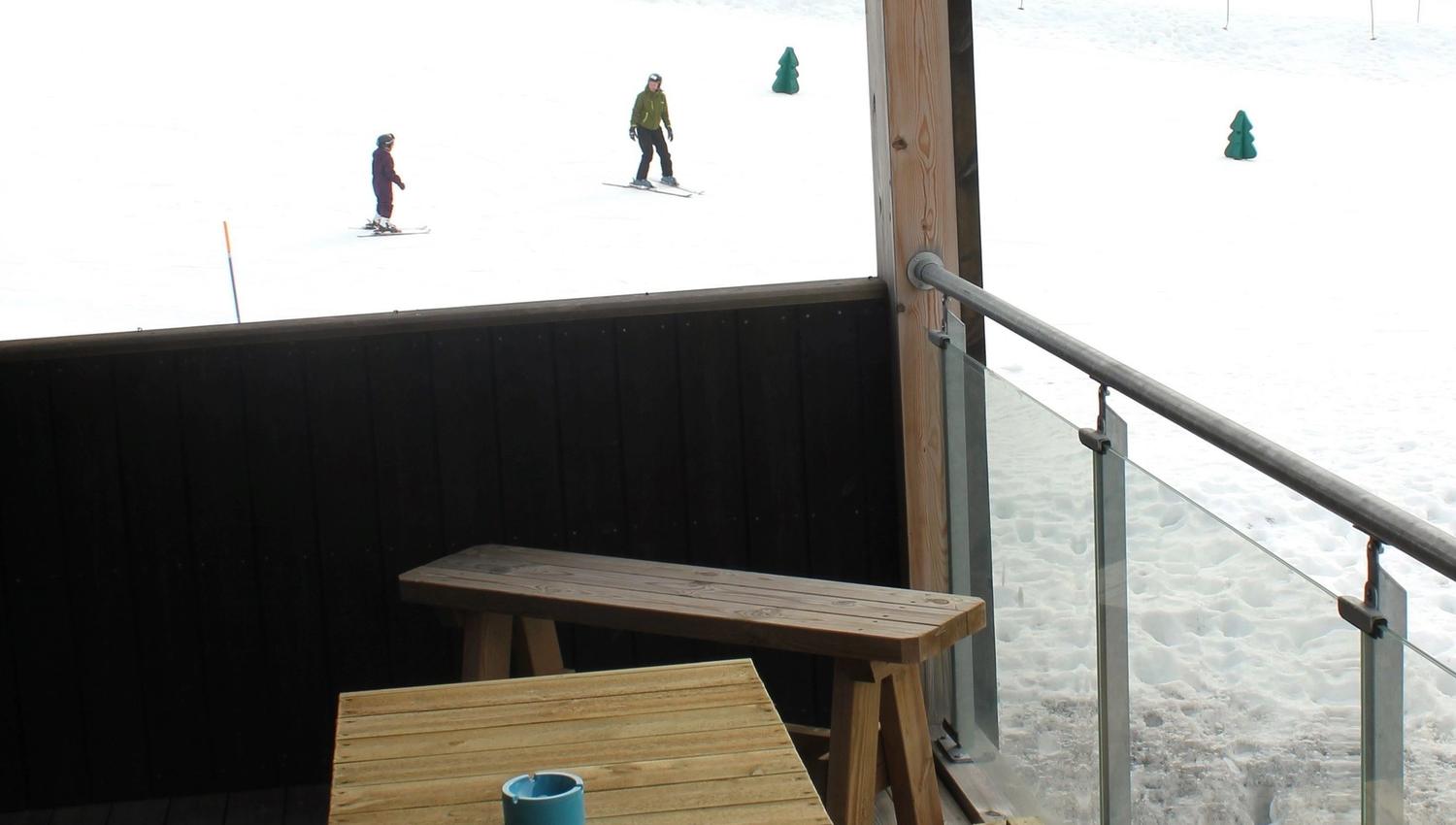 A table and benches on a terrace with snow and people skiing.