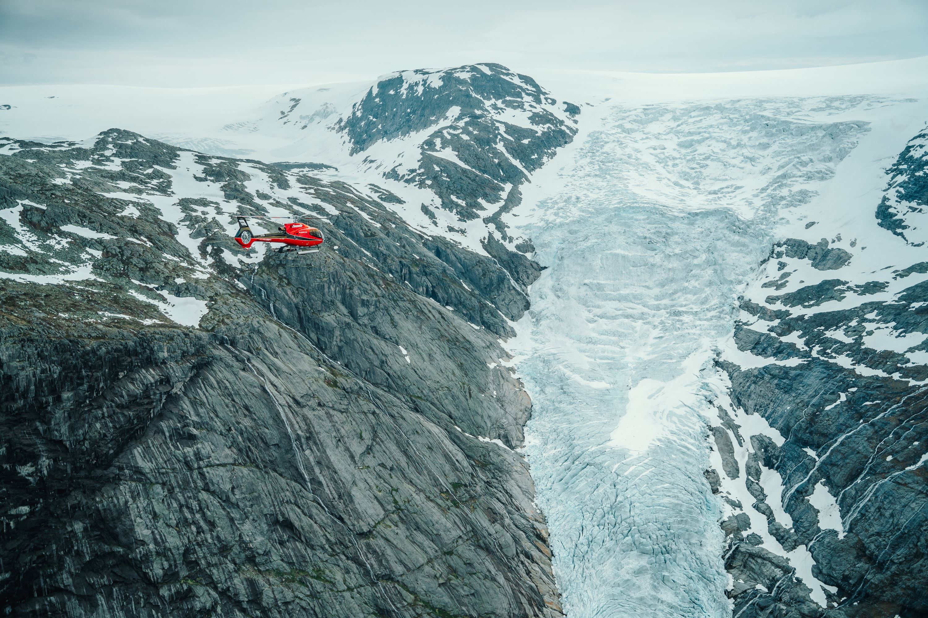 Fjord Helikopter  -Eit raudt helikopter flyg over bratte fjell og ein blåkvitt isbrelandskap under overskya himmel..