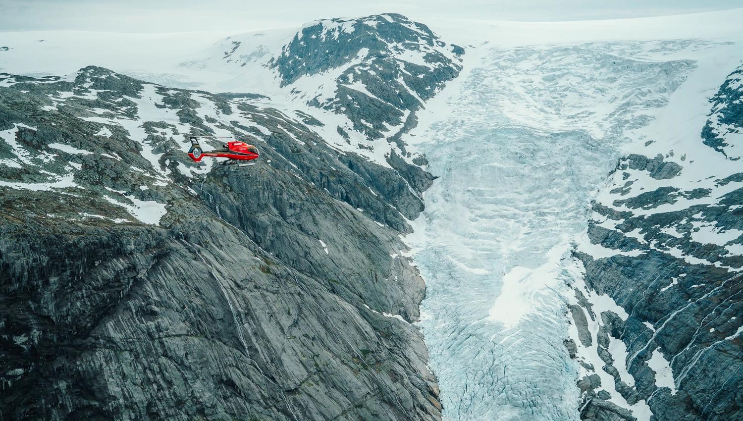 Fjord Helikopter -Eit raudt helikopter flyg over bratte fjell og ein blåkvitt isbrelandskap under overskya himmel..