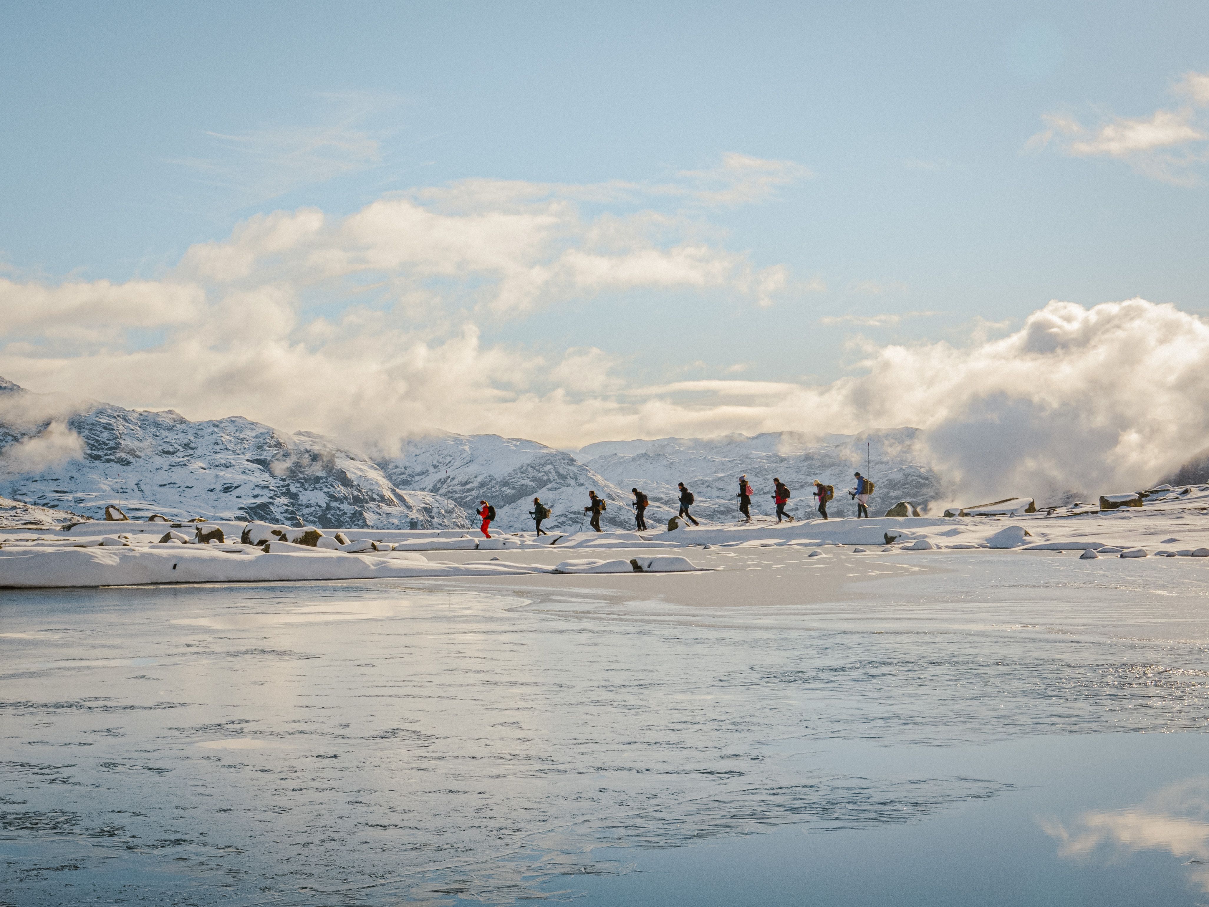 Winter group hiking across a frozen mountain plateau by a calm alpine lake near Trolltunga.