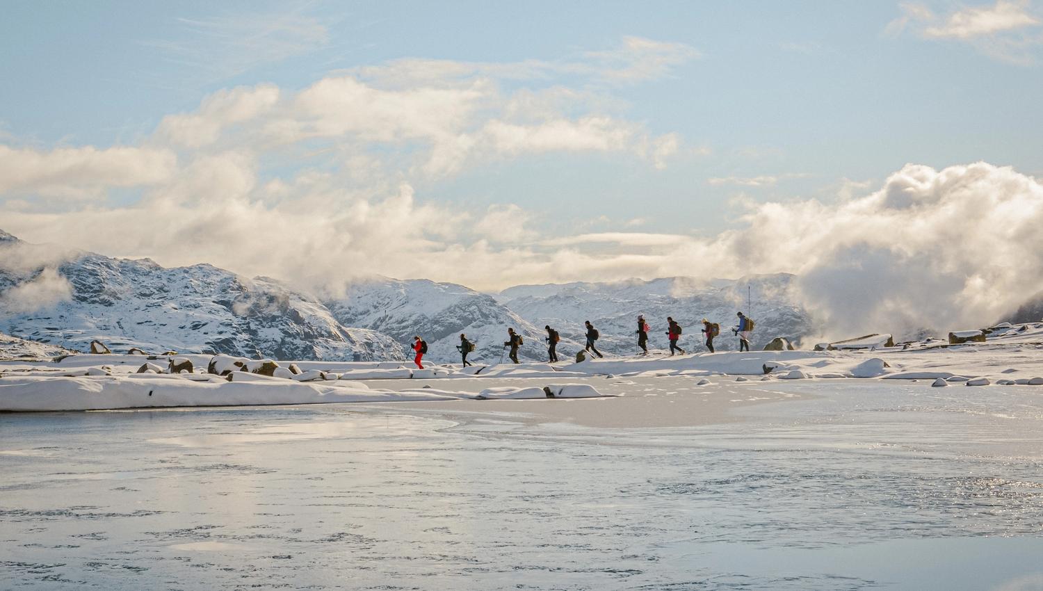 Winter group hiking across a frozen mountain plateau by a calm alpine lake near Trolltunga.