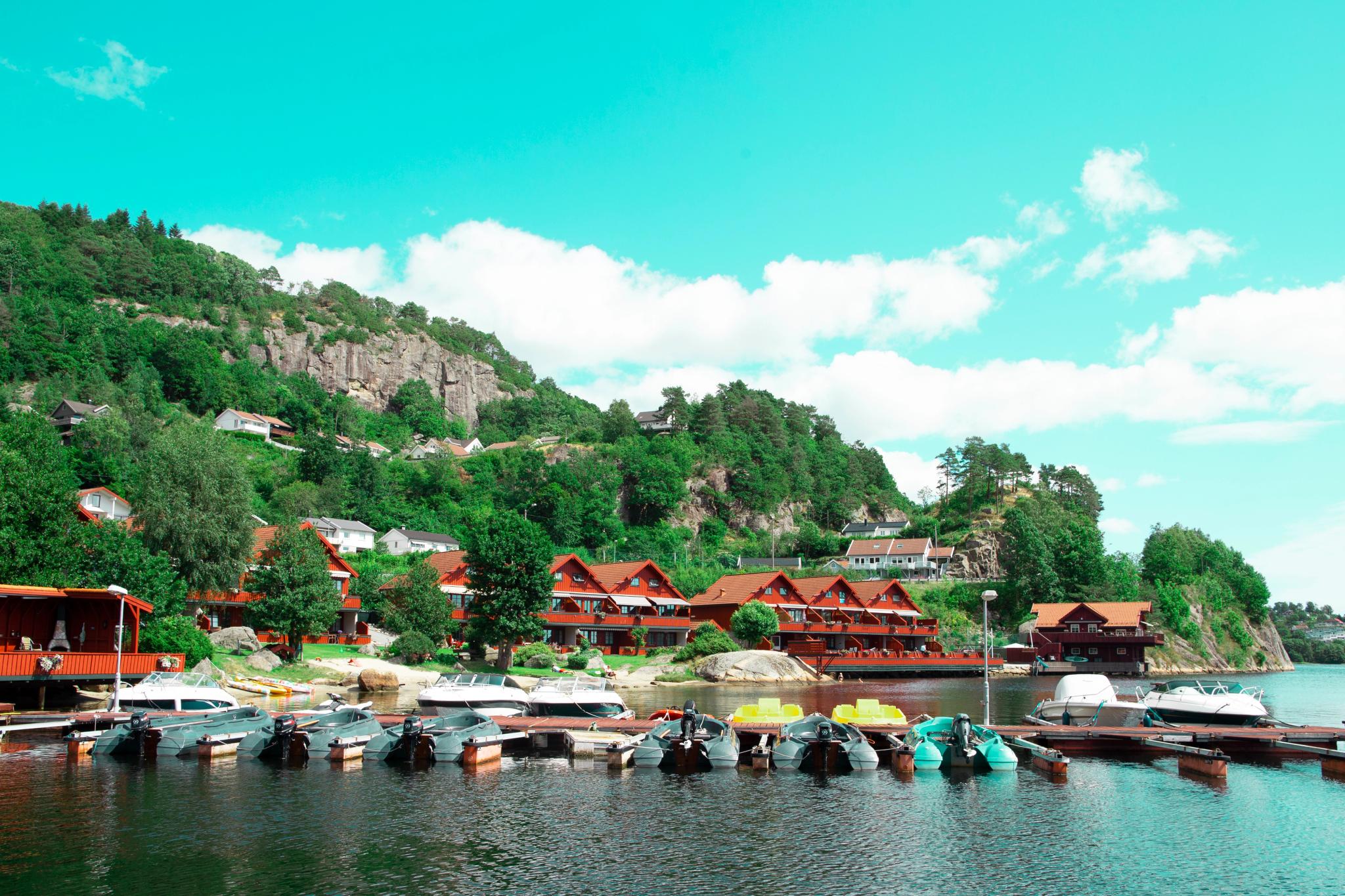 Rødbrune feriehus ved vannet, og en brygge som er fullt opp med båter. Skog og fjell bak feriehusene, blå himmel og litt skyer