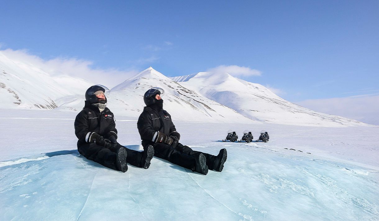 To personer i snøscooterutstyr som sitter på en isformasjon og slapper av i forgrunnen, med snøscootere parkert i et snødekt landskap i bakgrunnen