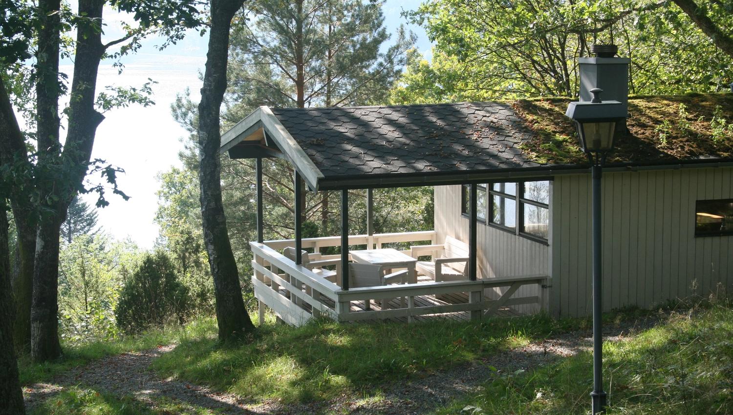 Hytte med terrasse omgitt av skog og utsikt mot fjorden.