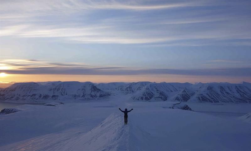 En person med utstrakte armer på en snødekt fjellrygg, med et mørkt fjellandskap og en solnedgang i bakgrunnen