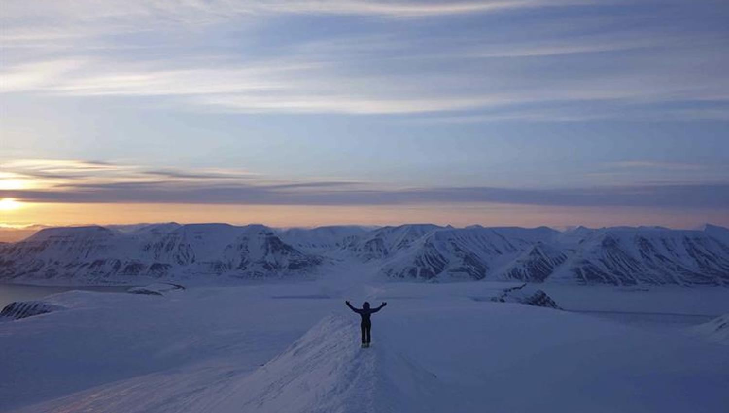 En person med utstrakte armer på en snødekt fjellrygg, med et mørkt fjellandskap og en solnedgang i bakgrunnen