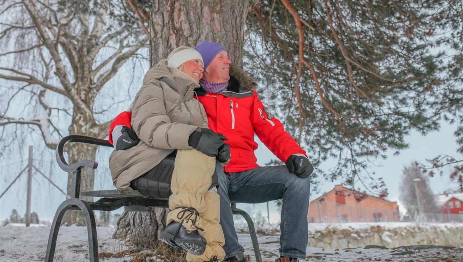 To personer sitter på en benk ved et tre i et snødekt område.