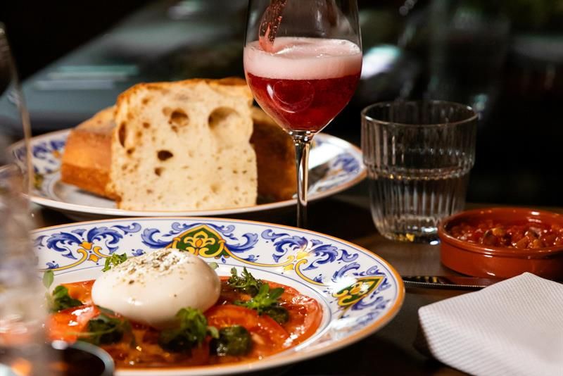 En flaske som heller en rosa drikk i et vinglass over et bord med burrata på tomat, brød, småretter og et glass vann.
