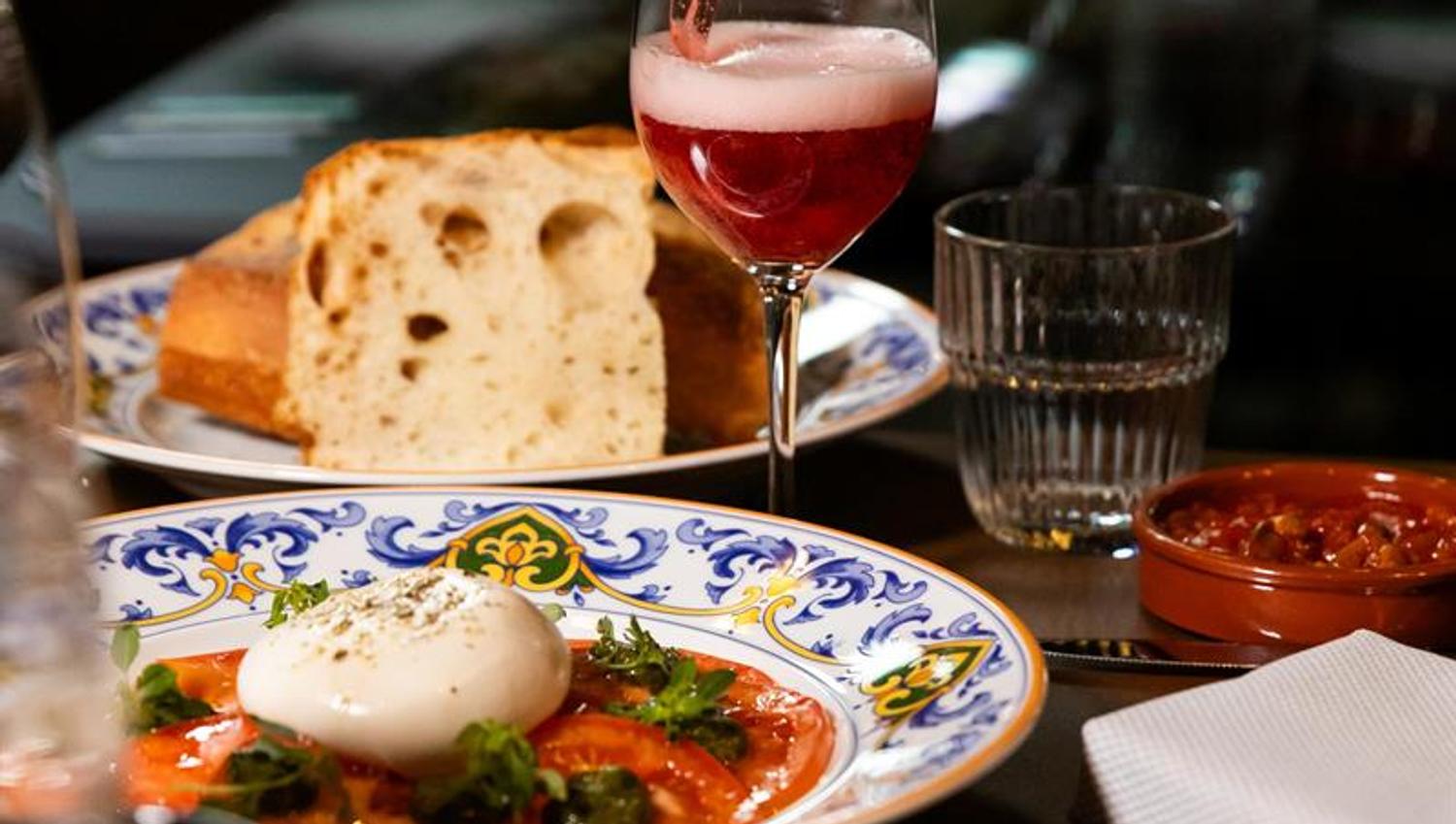 En flaske som heller en rosa drikk i et vinglass over et bord med burrata på tomat, brød, småretter og et glass vann.