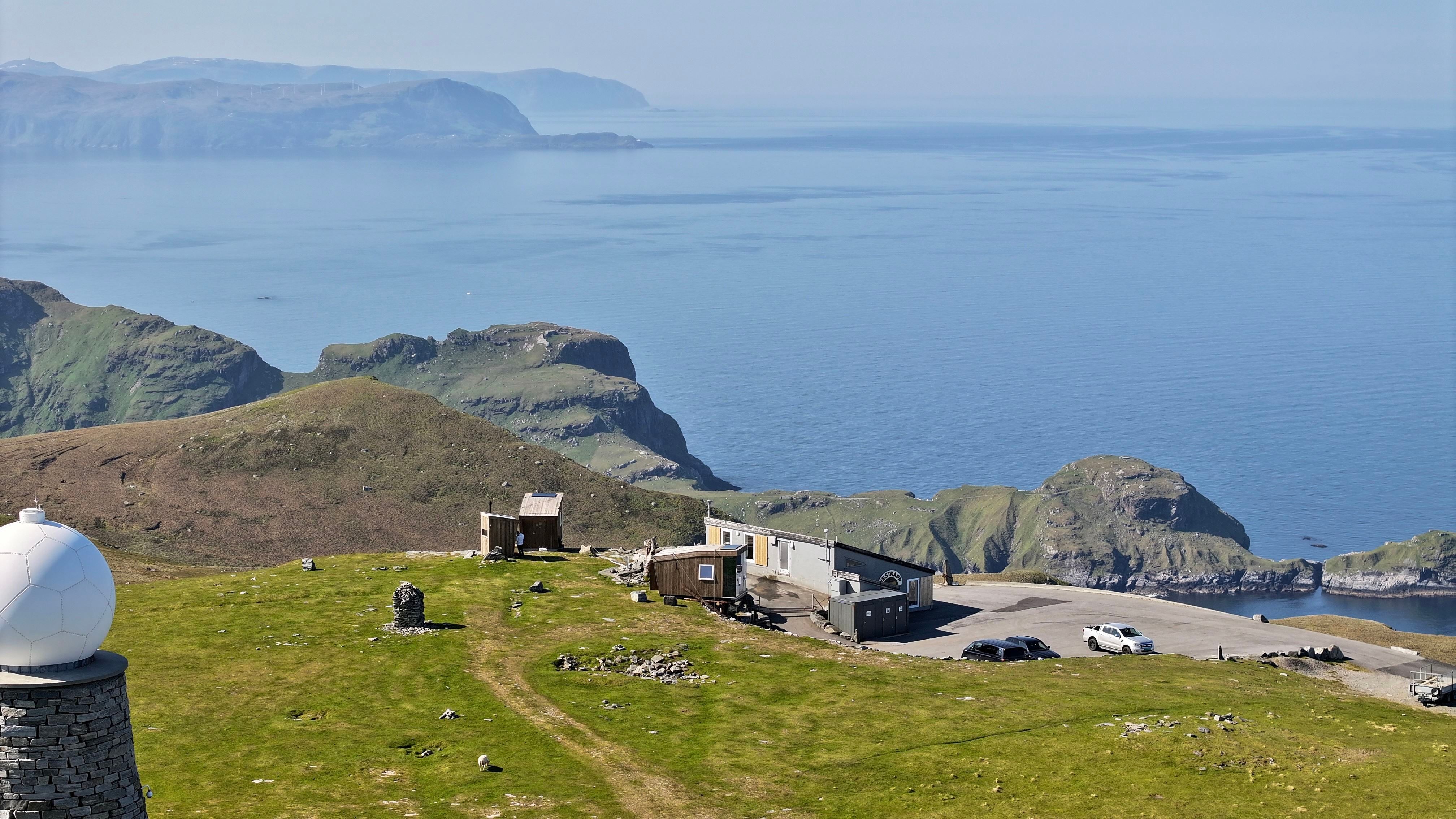 Grønt fjellplatå med små bygg og parkeringsplass, med bratte kystklipper og roleg blått hav i bakgrunnen..