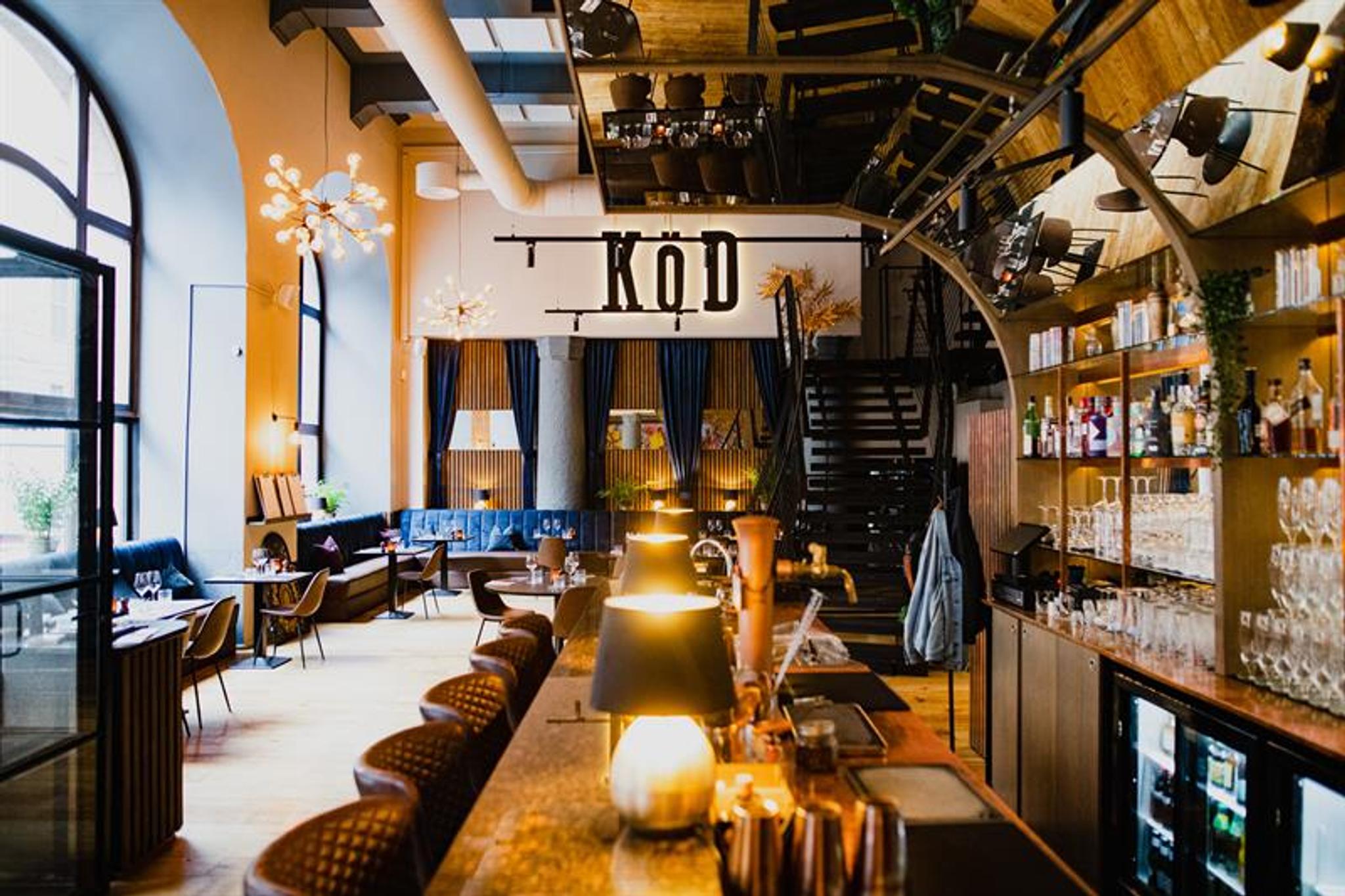 Restaurant interior with bar counter, bar stools, shelves with glasses and wine bottles, blue seating section along the wall and a high ceiling