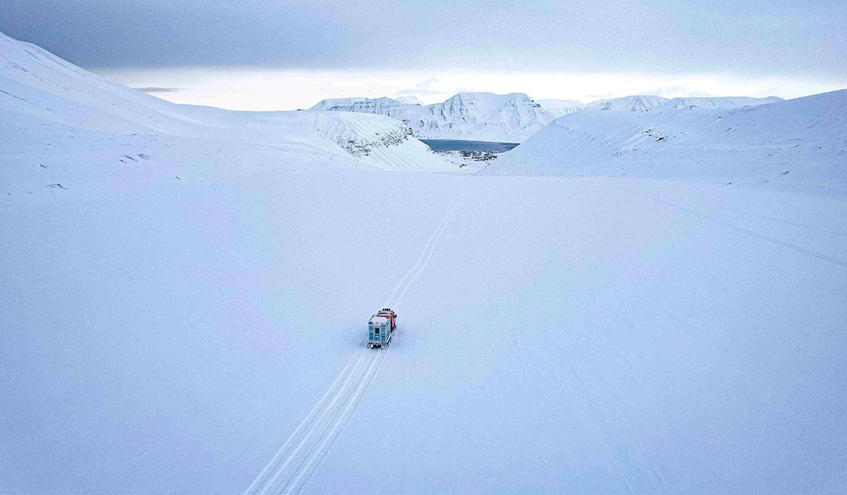 A snowcat on a glacier driving along tracks in the snow towards Longyearbyen in the background